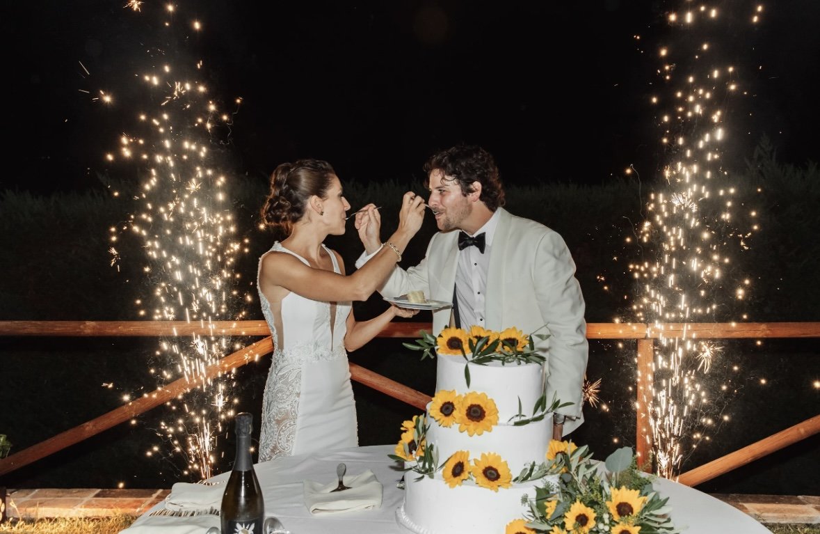 A bride and groom feeding each other cake at an outdoor wedding reception with fireworks in the background. The cake is decorated with sunflowers and a bottle of wine is on the table.