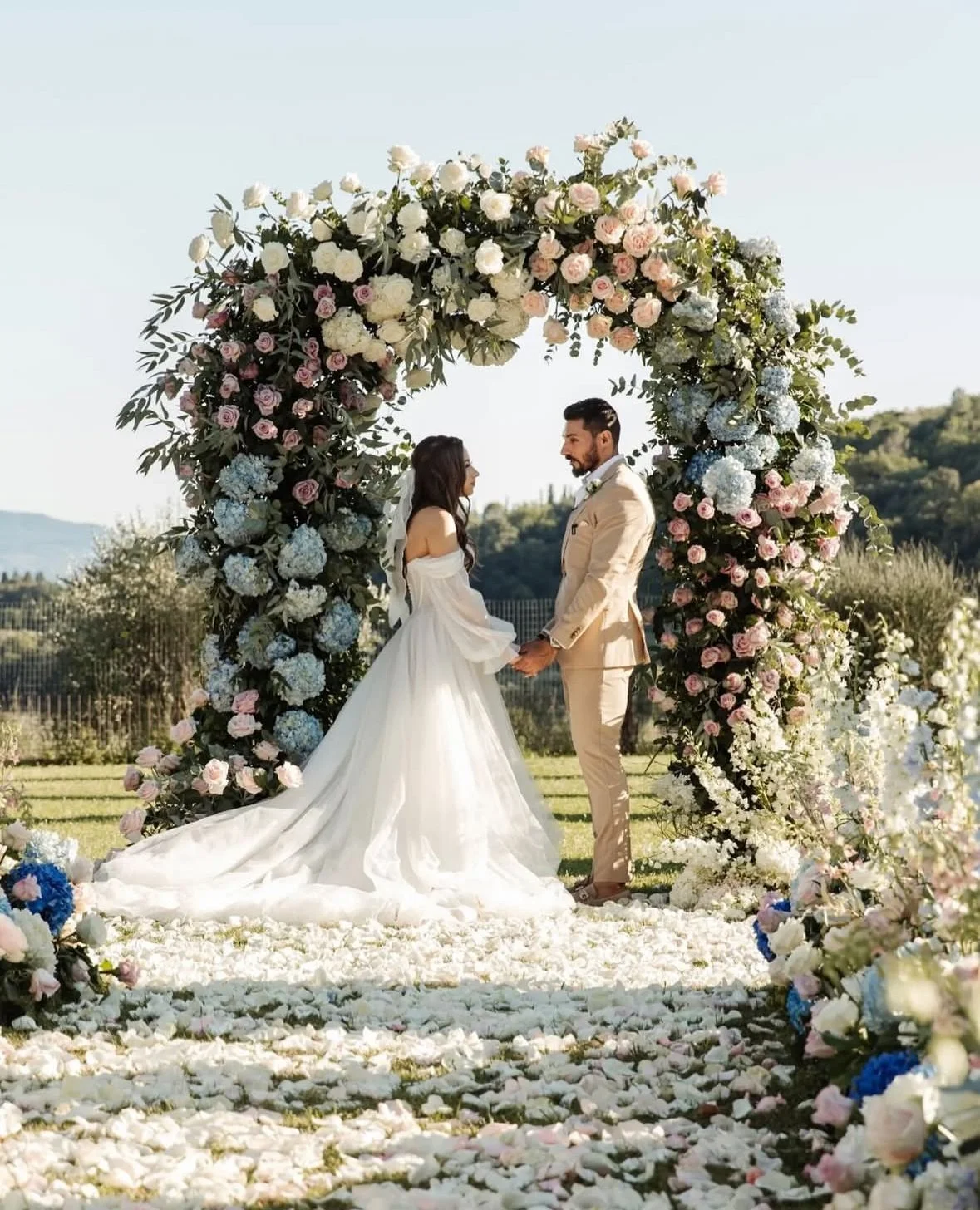 A bride and groom stand under a floral arch at an outdoor wedding ceremony. The arch is adorned with pink, white, and blue flowers, and blooms are scattered along the aisle. The bride wears a long white gown, and the groom is in a beige suit.