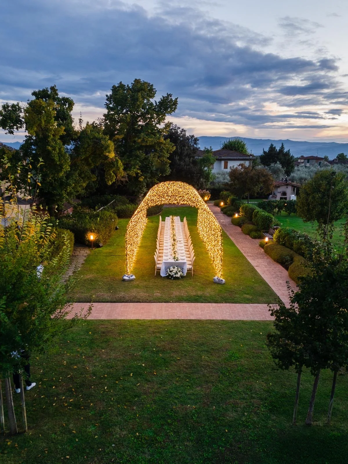 Outdoor wedding ceremony setup in a garden with a long aisle and floral archway illuminated by string lights, surrounded by trees and greenery under a cloudy sky.