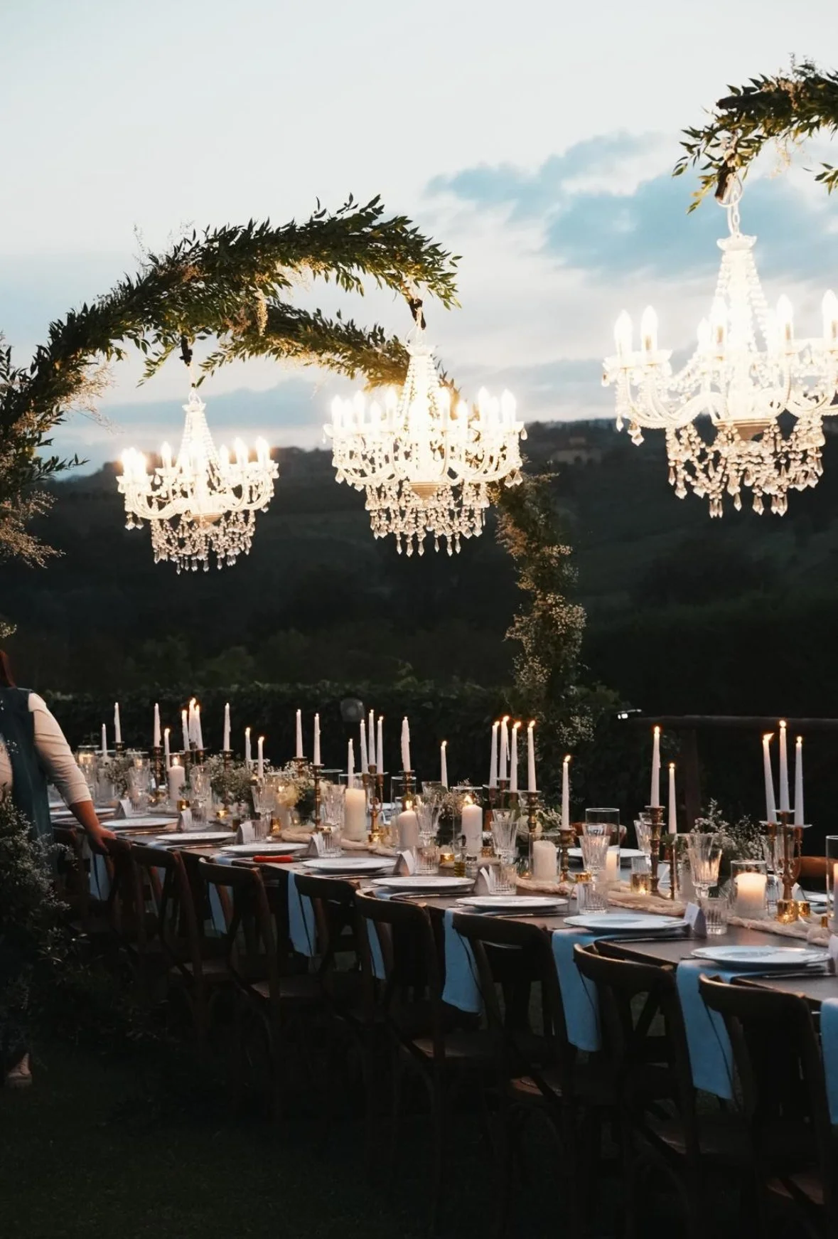 Outdoor dining setup with a long table, decorated with candles and chandeliers hanging overhead. The setting is surrounded by greenery and has a scenic background.