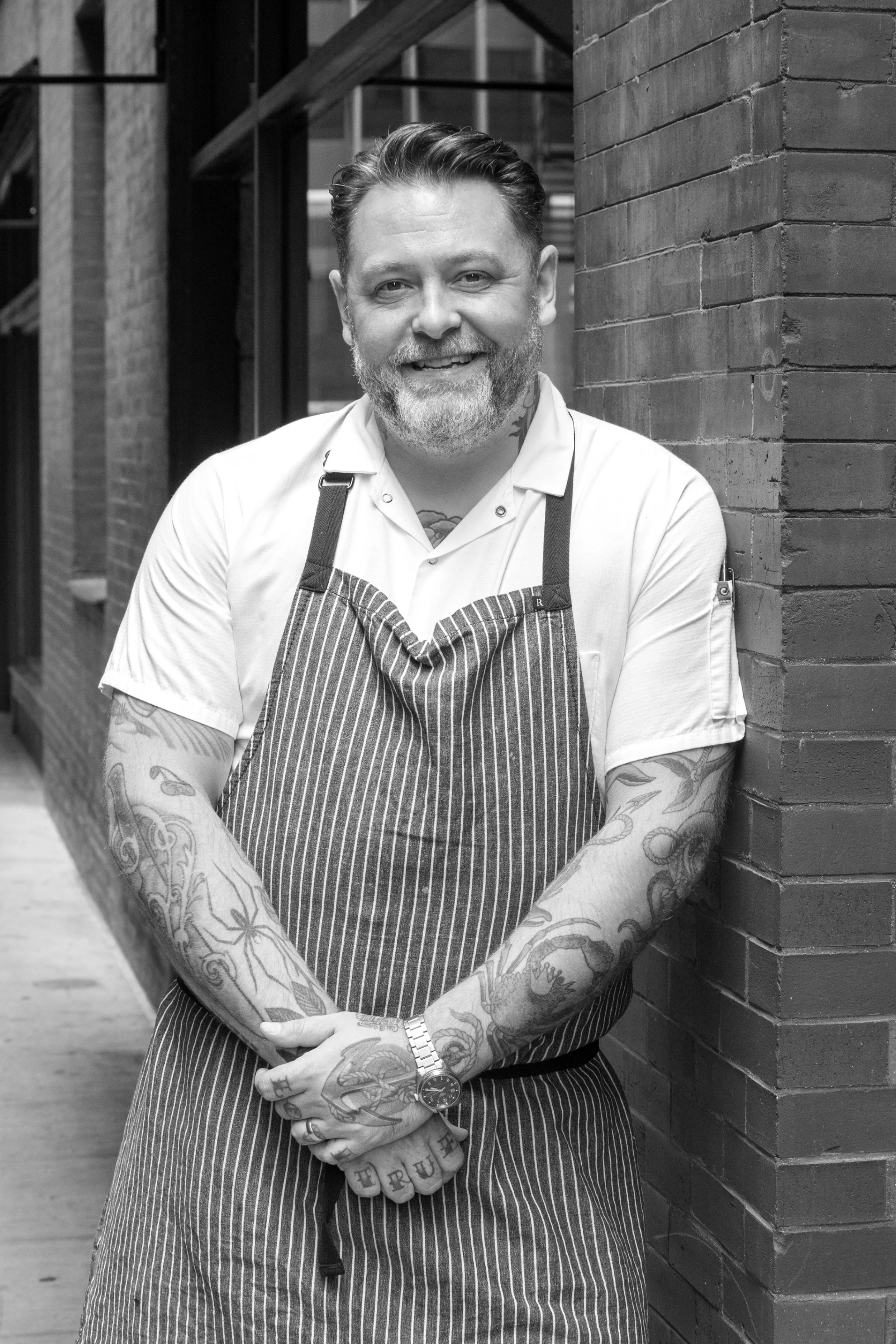 A smiling man with tattoos on his arms and hands, wearing a striped apron, standing outdoors next to a brick wall.