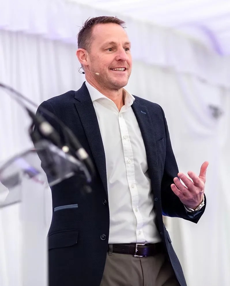 A man in a dark blazer and white shirt speaking at an event with a microphone, gesturing with his hand, in a bright indoor setting.