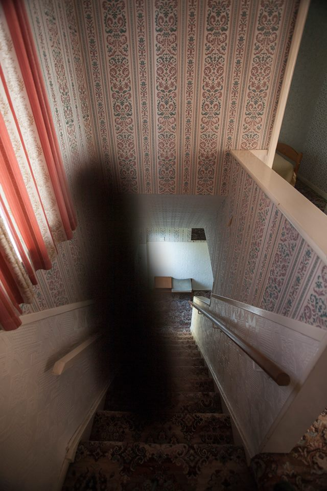 Staircase inside 30 East Drive in Pontefract, England, with patterned wallpaper and a large dark, shadowy figure appearing along the left side of the stairs.