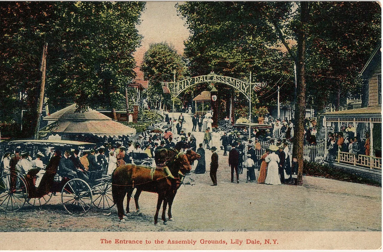 Vintage postcard showing the entrance to the Lily Dale Assembly in New York, with horse-drawn carriages and crowds of visitors gathering at the historic Spiritualist community in the early twentieth century.
