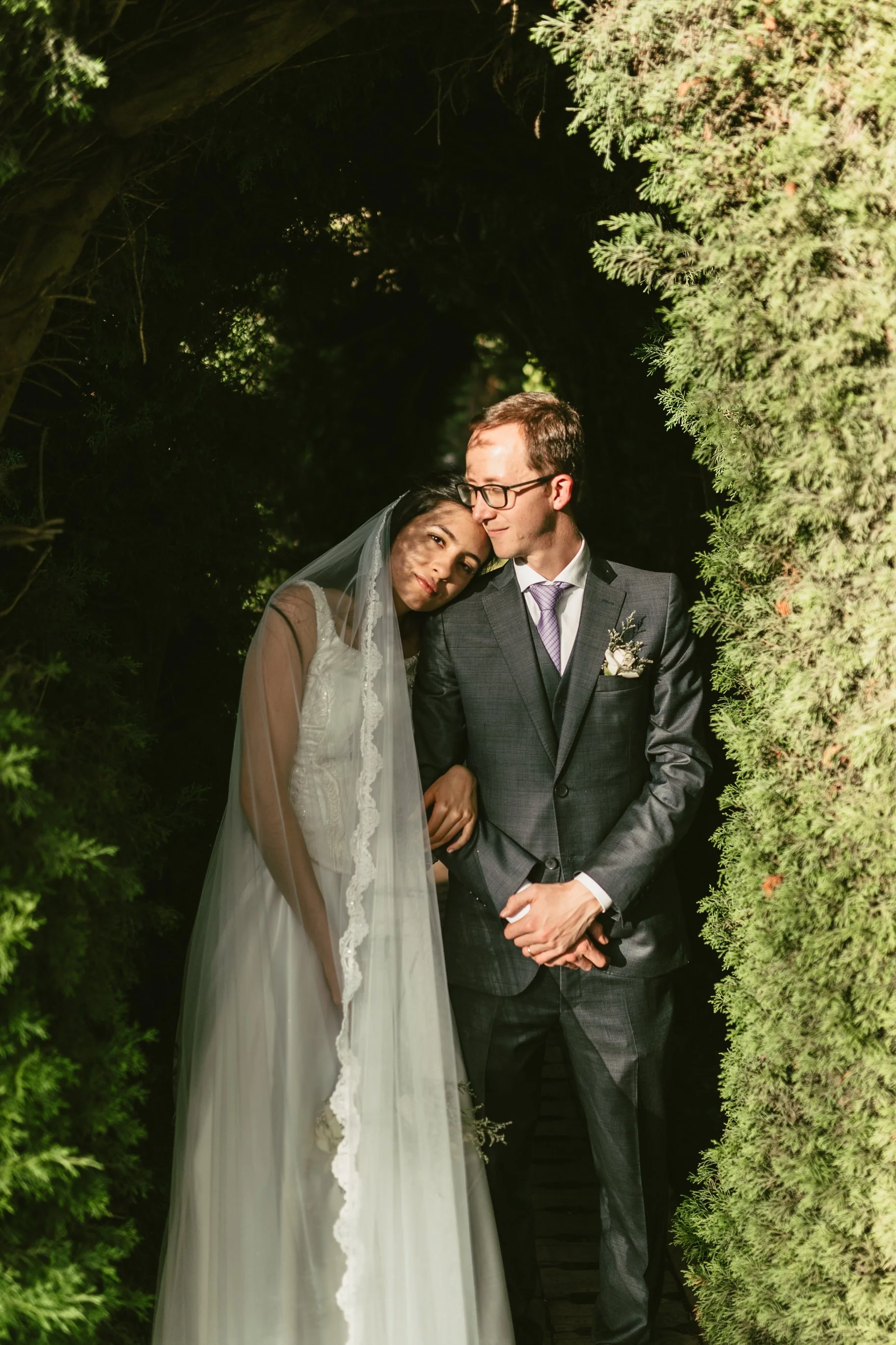 Una pareja de recién casados posando en un entorno natural rodeados de arbustos verdes, vistiendo el atuendo de boda, ella con vestido blanco y velo y él con traje gris y corbata