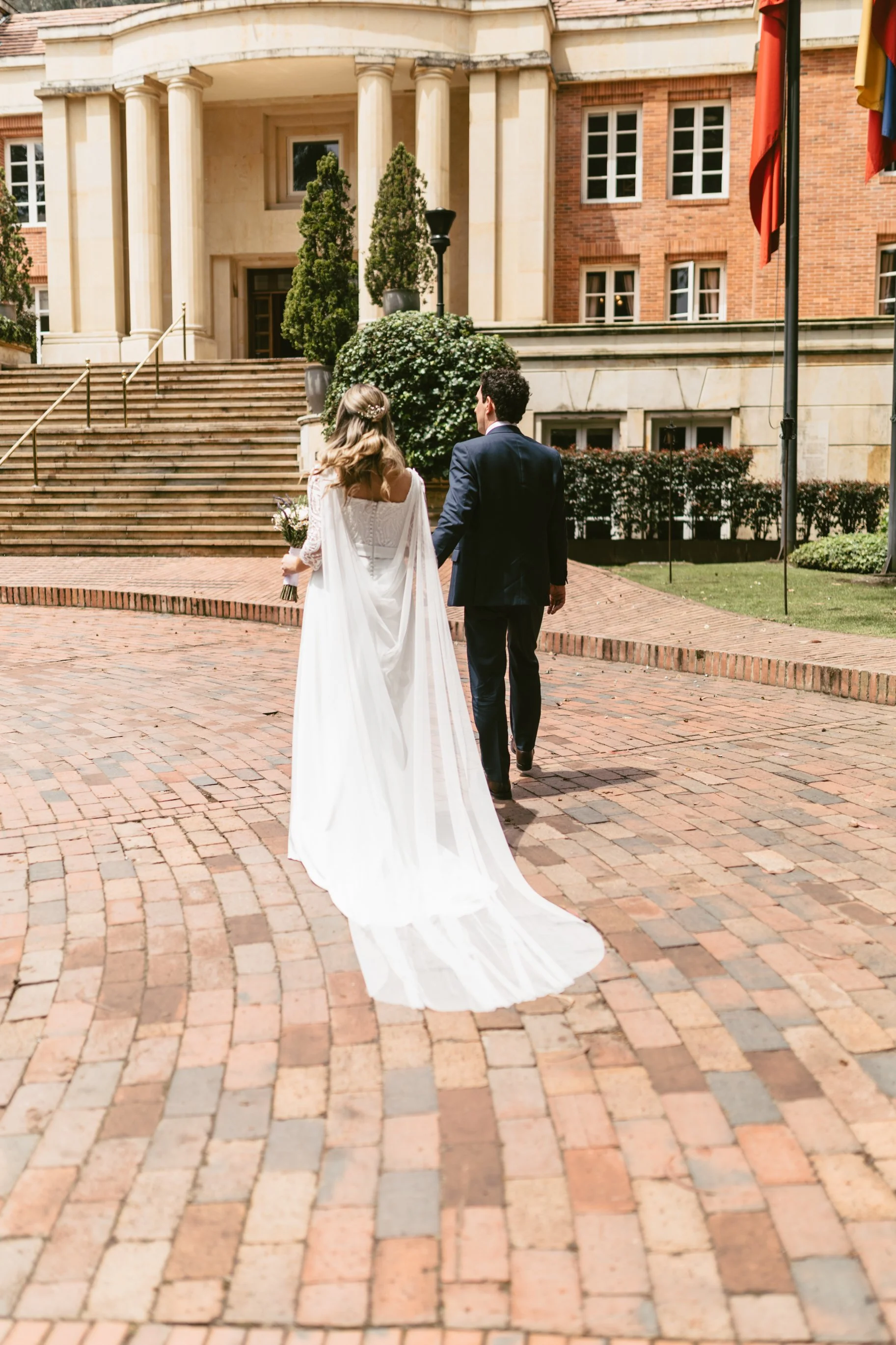 Pareja de novios caminando hacia un edificio de estilo clásico con columnas, la novia lleva vestido blanco y el novio traje oscuro, en un lugar exterior con adoquines y plantas ornamentales.