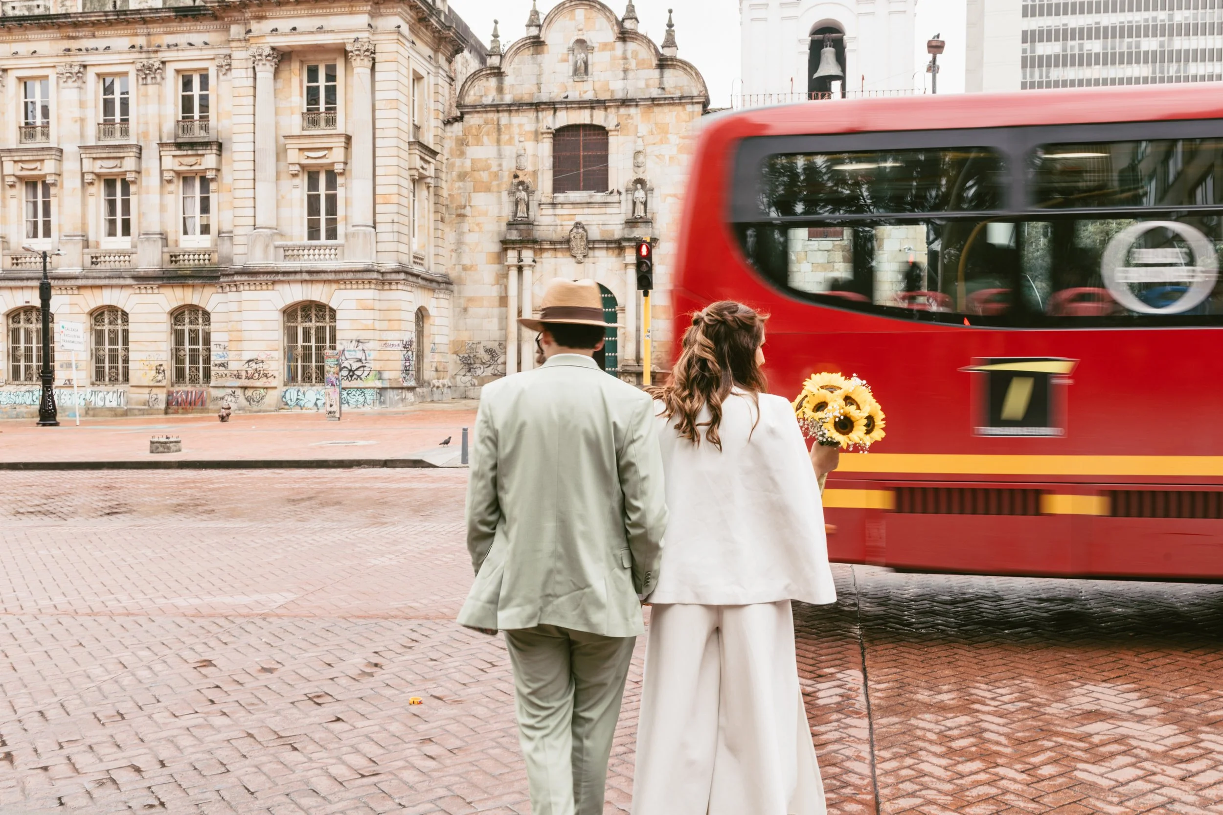 Una pareja vestida con ropa formal caminando en la calle, la mujer sostiene un ramo de girasoles y la escena incluye un autobús rojo en movimiento y un edificio antiguo de arquitectura clásica en el fondo.
