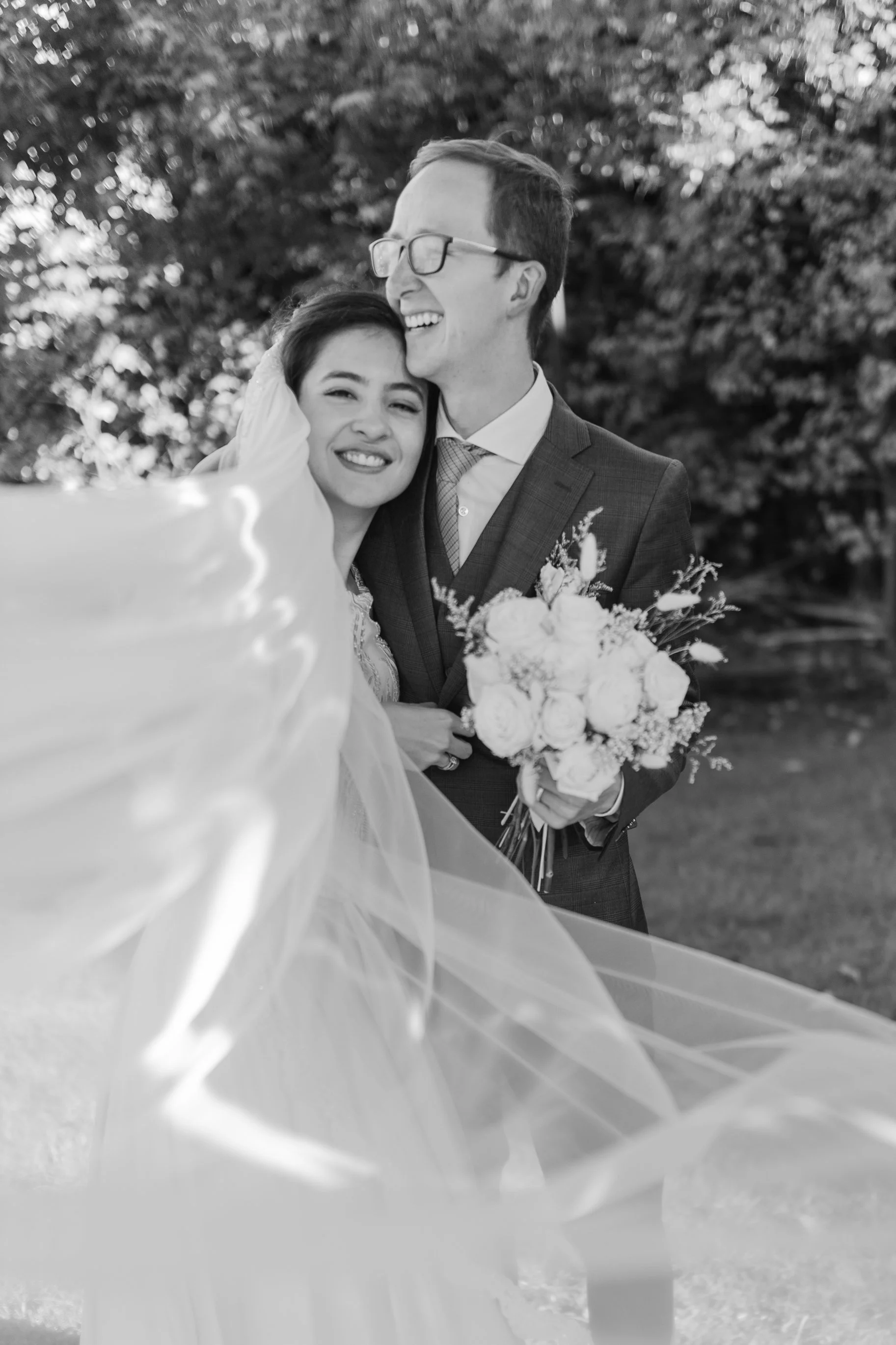 Una pareja de recién casados en un jardín, ella con velo y él con un ramo de flores, sonriendo y abrazándose, en una fotografía en blanco y negro.