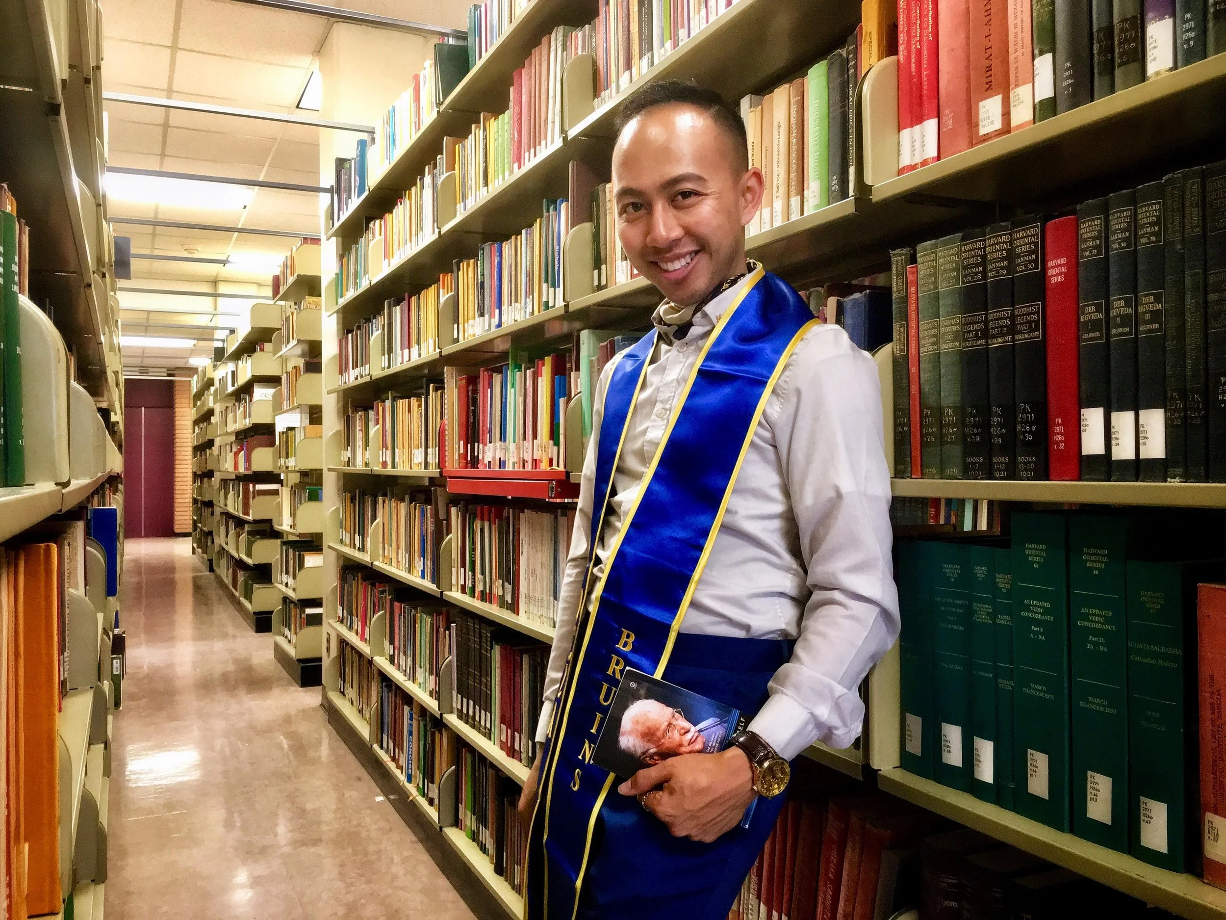 A young man in a graduation gown and sash standing between bookshelves in a library, holding a book with a photo of an elderly man on the cover.