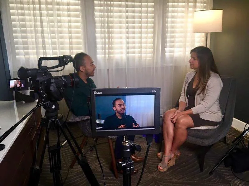 A woman being interviewed on camera in a room with white curtains, a lamp, and a grey chair.