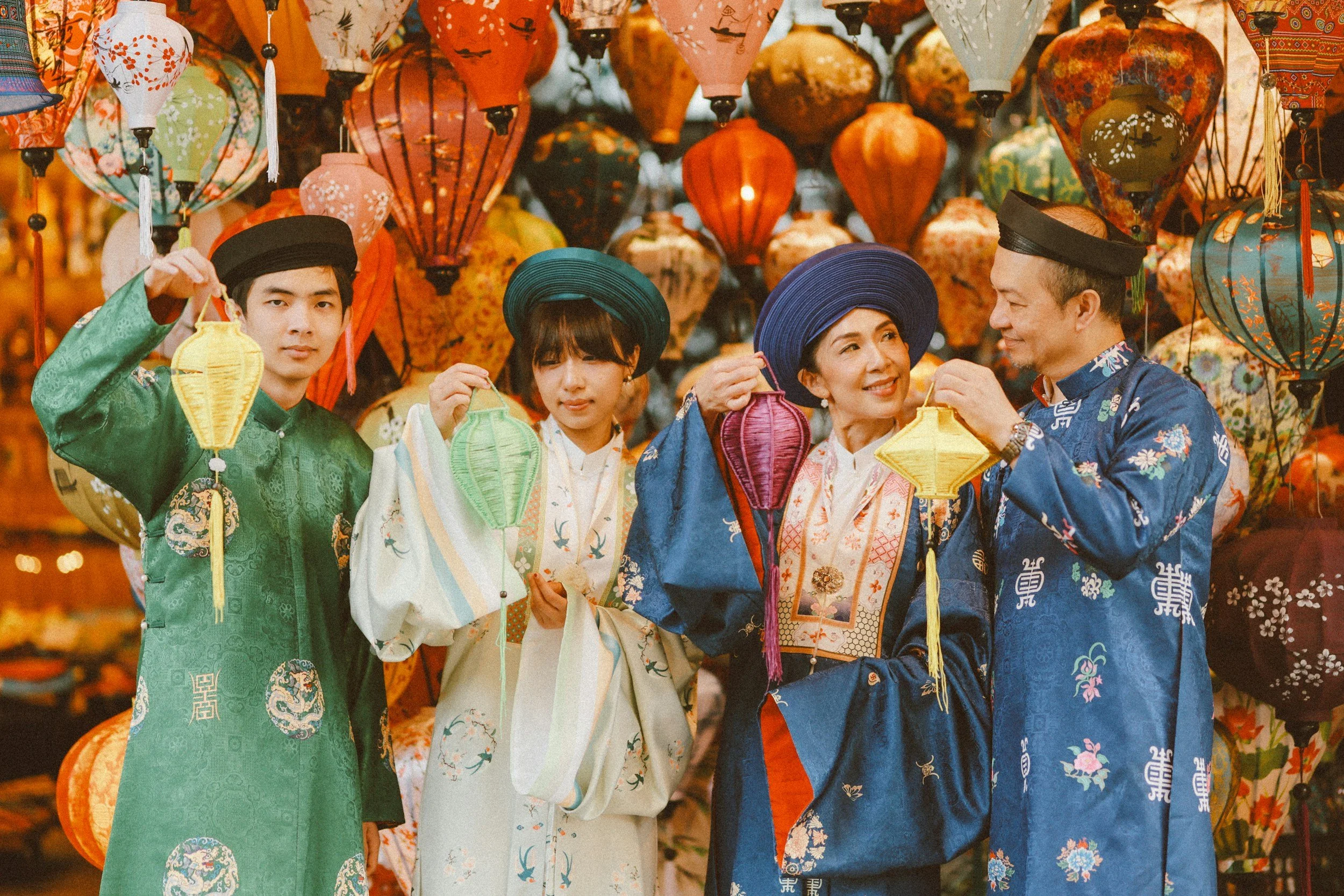 Group of four people wearing traditional Asian clothing and holding colorful lanterns, standing in front of a background filled with hanging lanterns of various shapes, sizes, and vibrant colors.