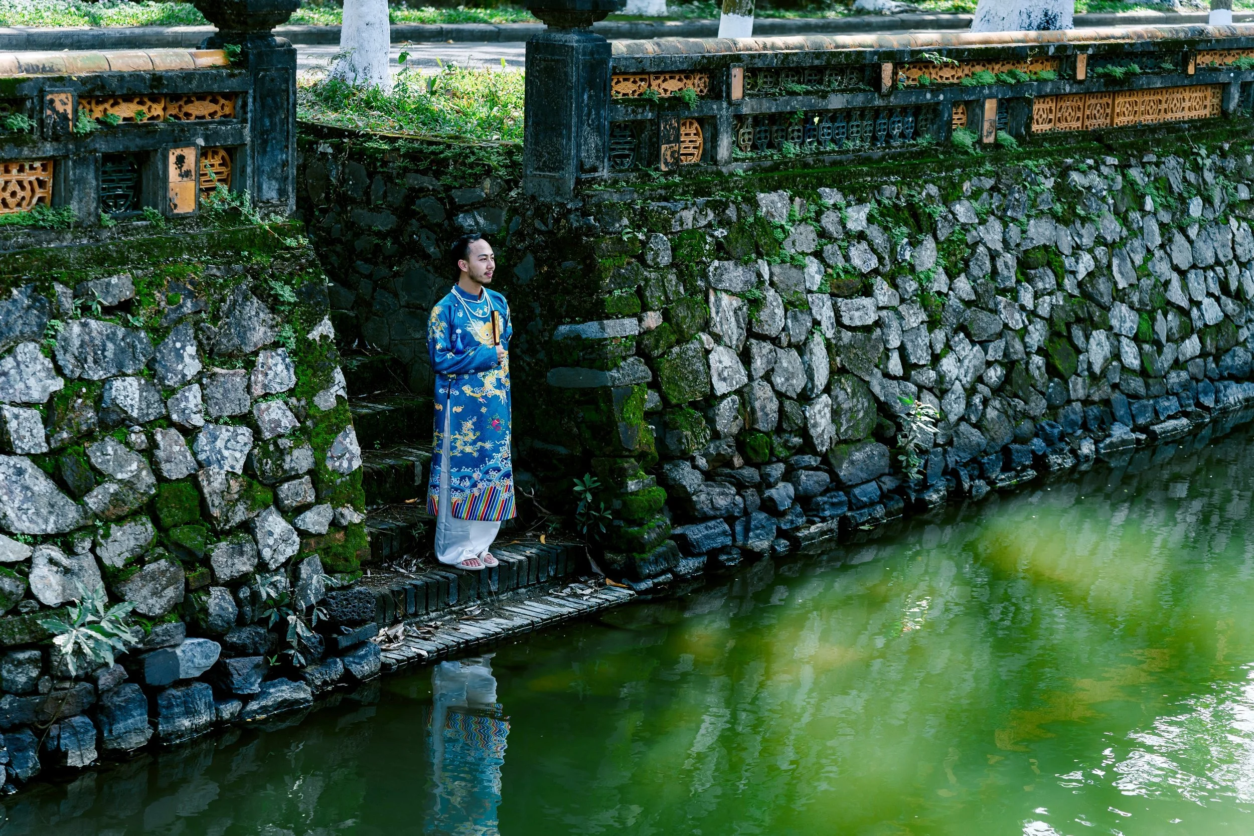 A man dressed in a traditional bright blue robe with intricate floral patterns stands on a small wooden bridge over a greenish river, surrounded by stone and moss-covered walls and lush trees.