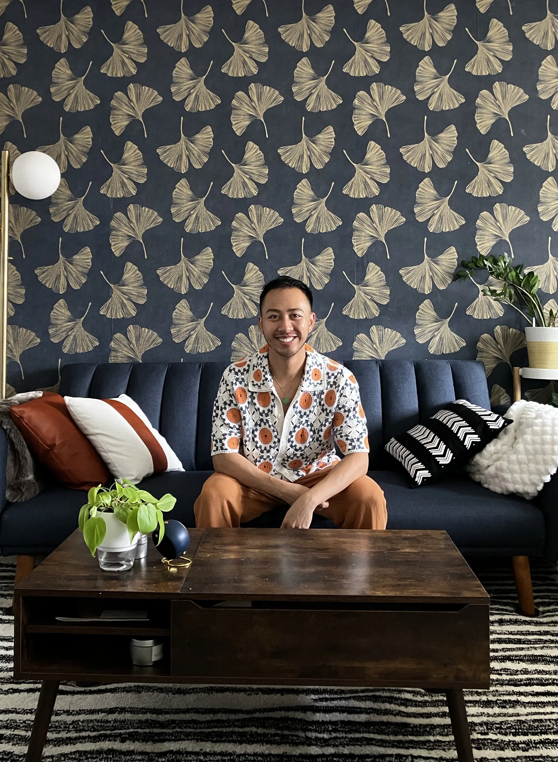 A smiling man with short dark hair sits on a dark gray sofa in a living room. He is wearing a white shirt with a colorful circular pattern and brown pants. Behind him is a dark blue wall with a pattern of large, golden ginkgo leaves. To his left, there are four cushions—two are white with a brown stripe, one is black and white striped, and the last is fluffy white. To his right, a white pot with a green plant and a led light are visible. There is a dark wooden coffee table with a plant, a black object, and a pair of glasses in front of him. The room has a striped black and white rug.