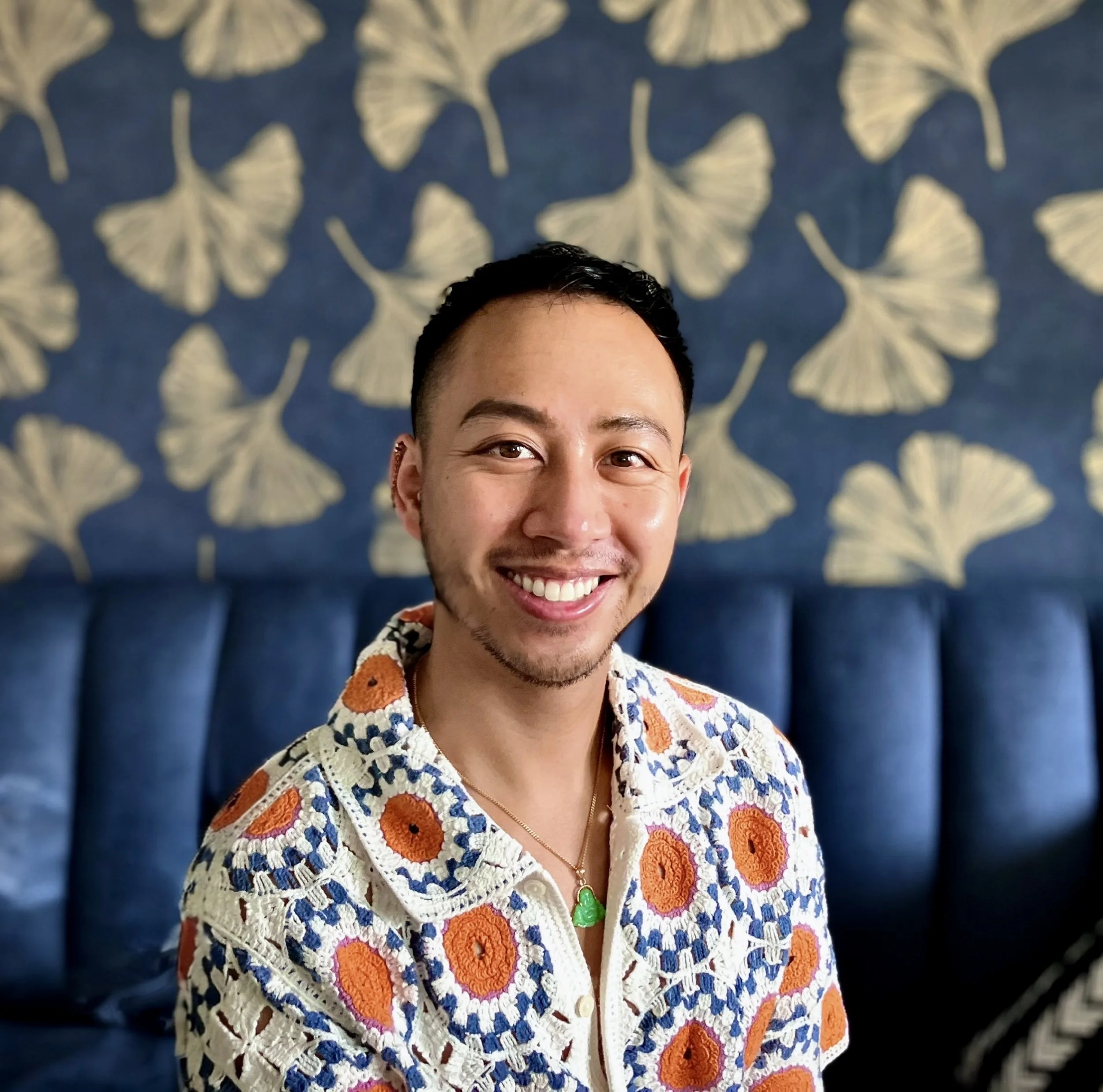 Close-up of a smiling man with short dark hair and a patterned shirt, standing in front of a blue wall with a white floral pattern.