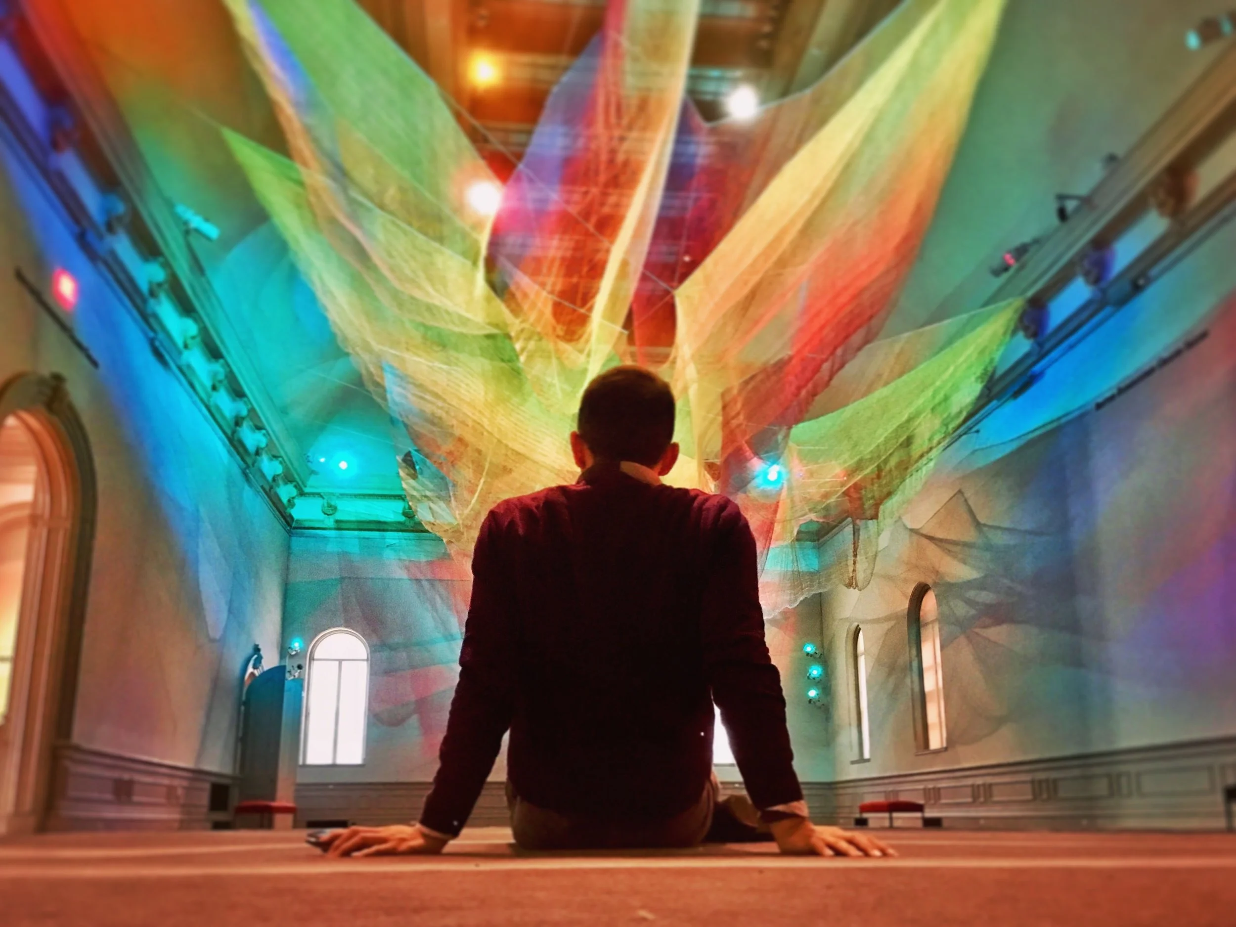 A person sitting on a wooden floor, facing away, in a large room with vibrant, colorful laser light projections on the ceiling and walls.