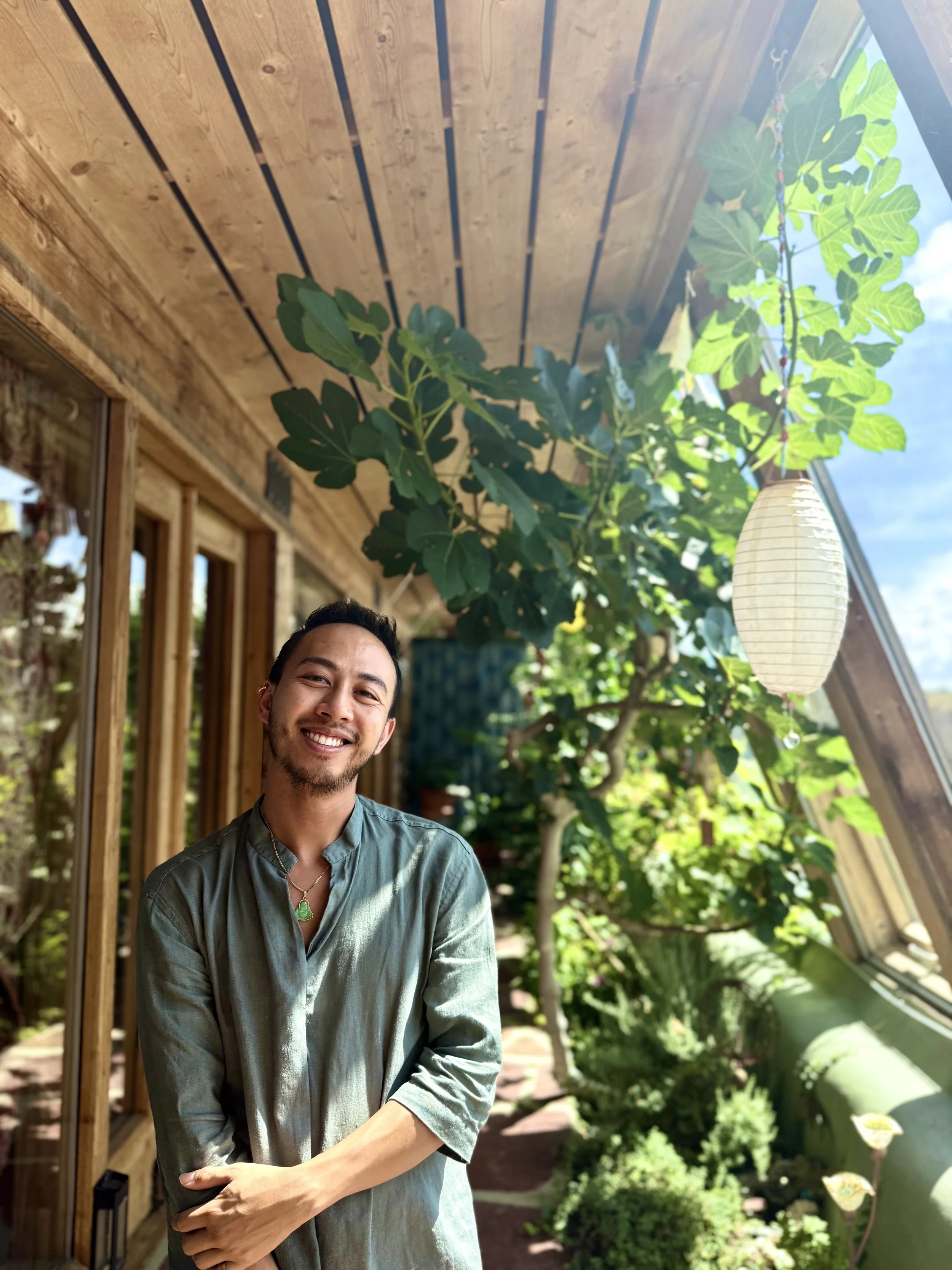 A smiling man with dark hair and a beard standing on a balcony or patio area with wooden beams and lush green plants, including a large leafy plant and a paper lantern hanging from the ceiling, in a sunny outdoor setting.