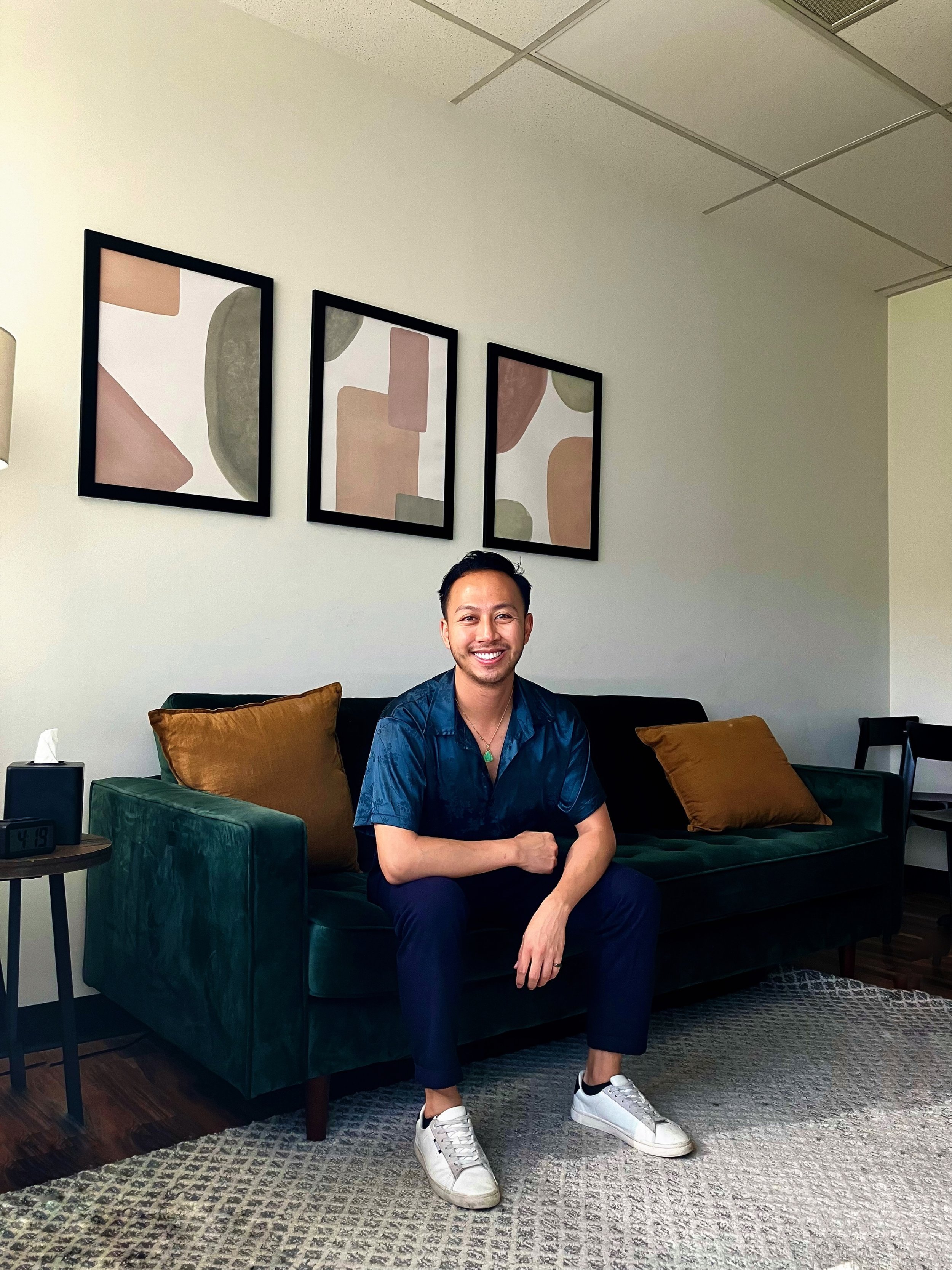 A smiling man with short dark hair, wearing a blue shirt, black pants, and white sneakers, sitting on a dark green velvet sofa in a room with white walls, three framed abstract art pieces, a small side table with a clock showing 4:19, a tissue box, and a rug on the floor.