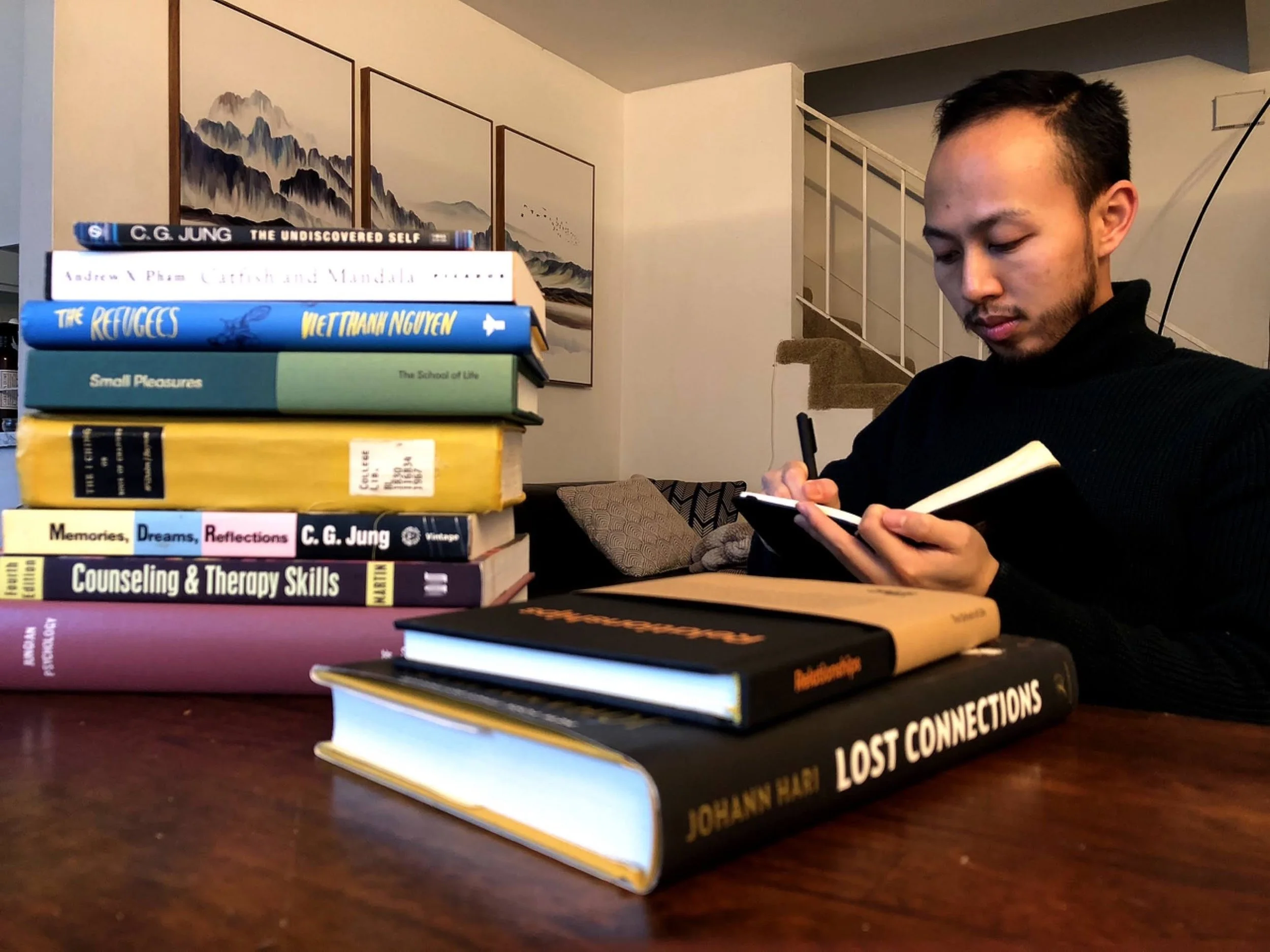 A person with short dark hair and facial hair, wearing a dark turtleneck, is sitting at a table behind a stack of books. The books include titles such as "The Uncovered Self," "The Refugees," "Small Pleasures,"