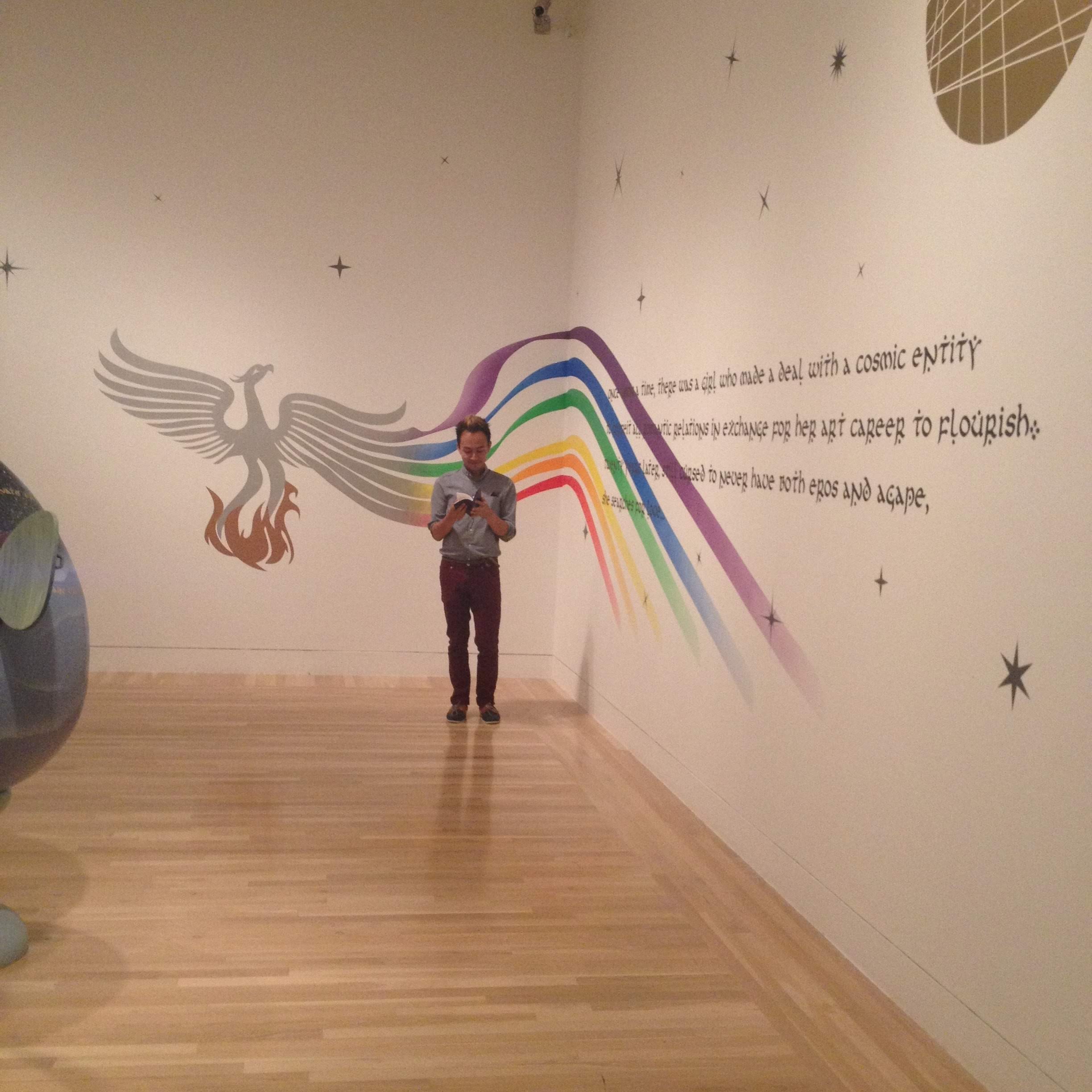 A person standing in front of a large wall mural featuring a rainbow, a phoenix, stars, and moon designs, with text about artistic exchange and cosmic identity.