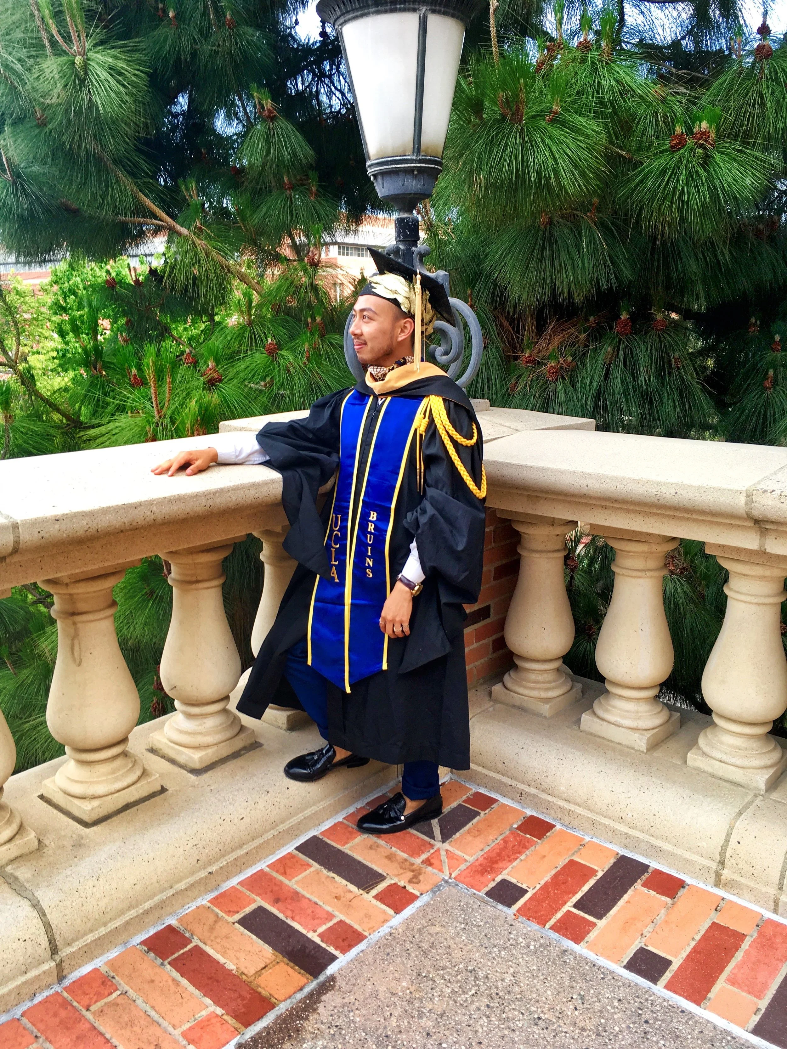 Graduation graduate wearing a cap and gown with a yellow sash, standing on a brick and concrete patio with ornate balustrade, green trees in the background.