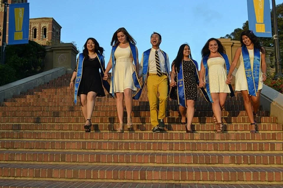 Six graduates wearing blue and gold robes holding hands and walking down outdoor brick stairs, smiling and celebrating graduation.