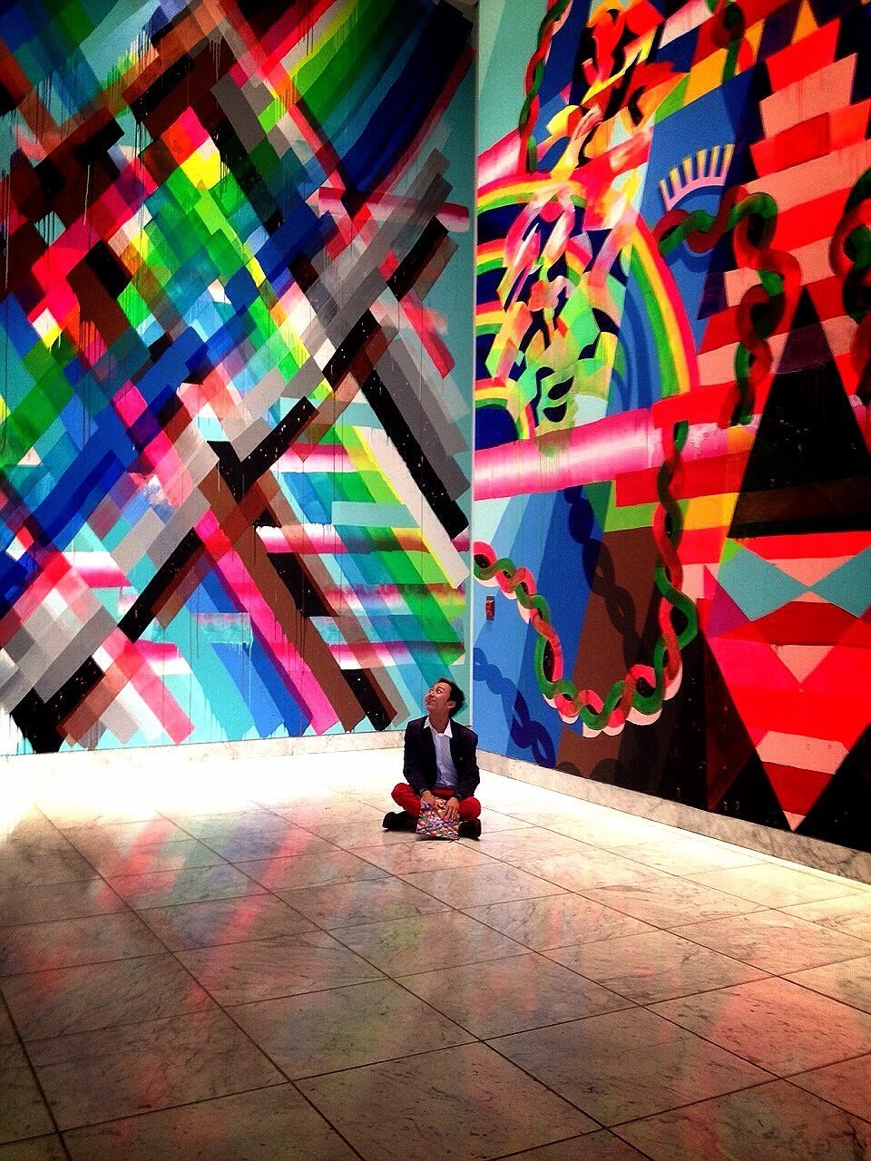 A person sitting on the floor in front of vibrant, abstract, colorful wall art with geometric shapes and patterns at an art gallery.