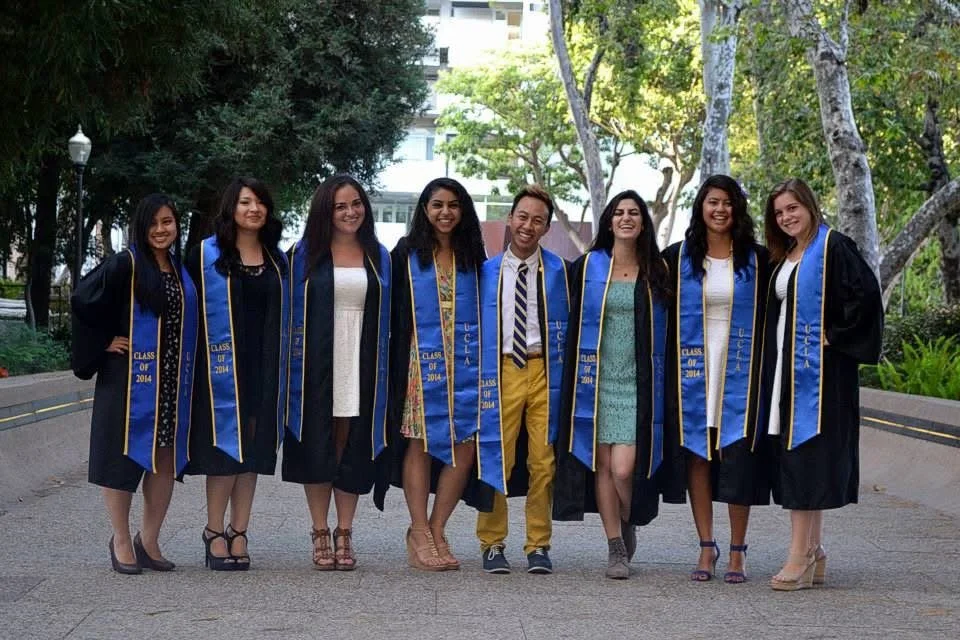 Group of eight diverse young adults in graduation caps and gowns, smiling, celebrating graduation outdoors.