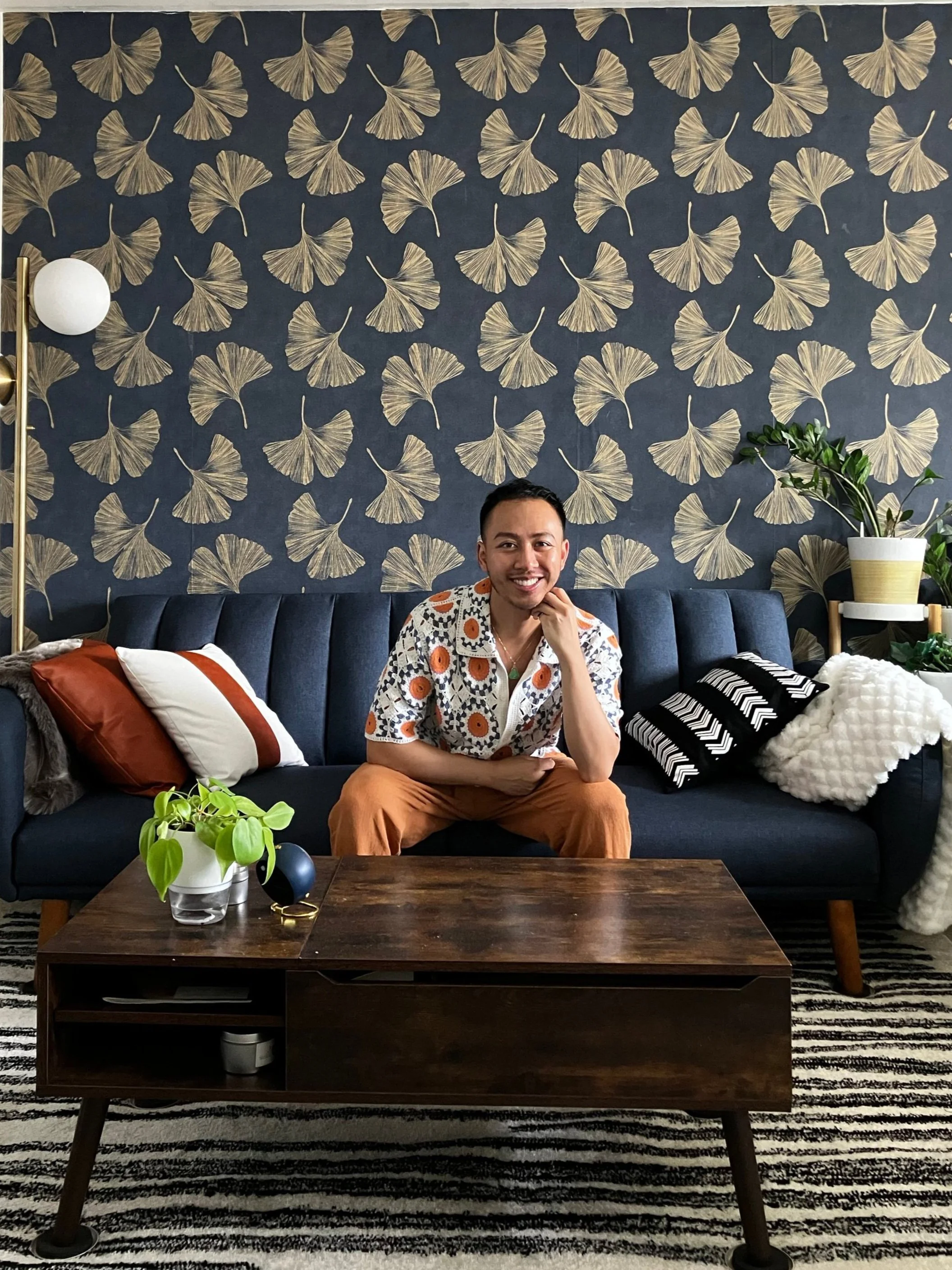 A man smiling and sitting on a dark blue sofa in a living room with a floral wallpaper, a white and orange pillow, and a black and white striped pillow, in front of a wooden coffee table with plants, a blue vase, and glasses, and a tall lamp with a white globe shade and a white and yellow pot plant.