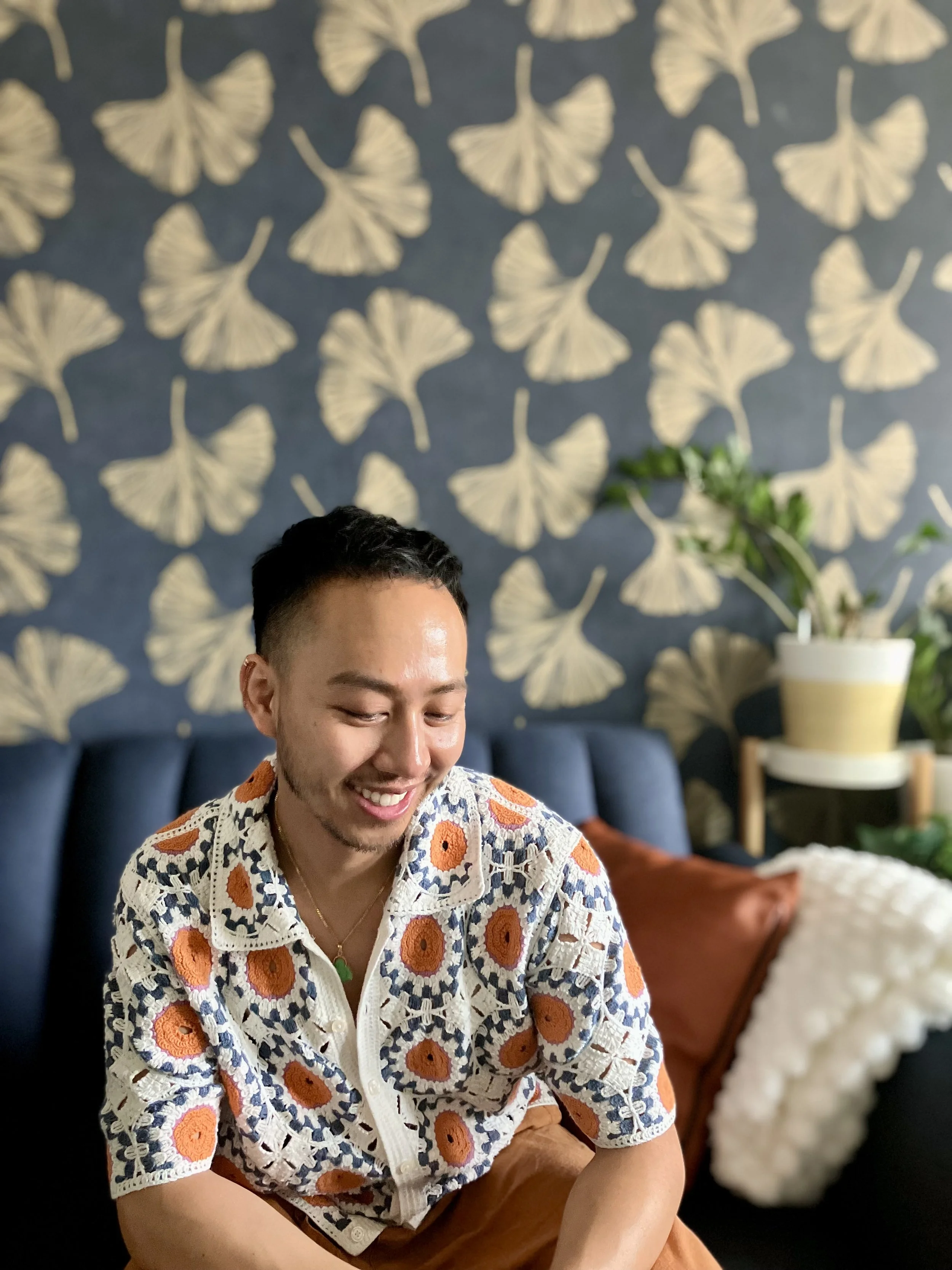 A smiling man with short dark hair and light skin sitting in a room with a dark blue wall that has a cream-colored ginkgo leaf pattern. He is wearing a white crochet shirt with orange, navy, and cream floral designs, and brown pants. There is a potted plant on a side table and a floral arrangement in the background.