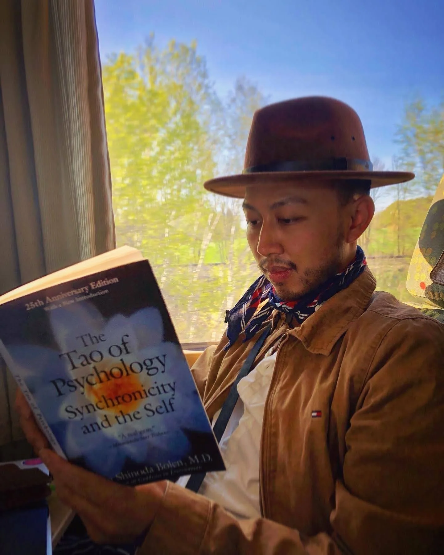 A man sitting on a train or bus reading a book titled 'The Tao of Psychology.' He is wearing a brown hat, a brown jacket, a white shirt, and a scarf. The background shows trees and clear skies outside the window.