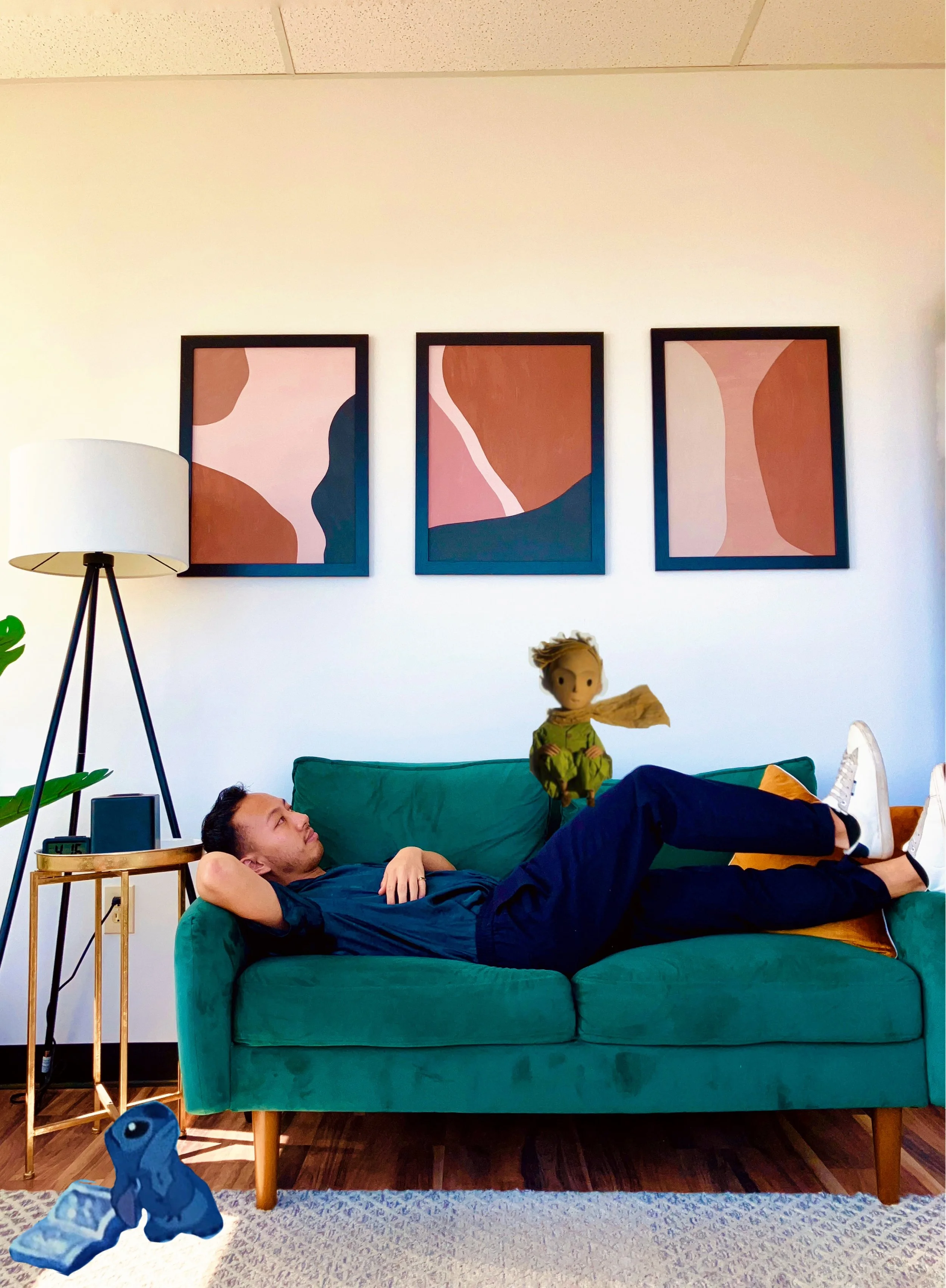 A man lying on a teal sofa in a living room with artwork on the wall, a floor lamp, a side table with a digital clock, and a plush toy on the floor.