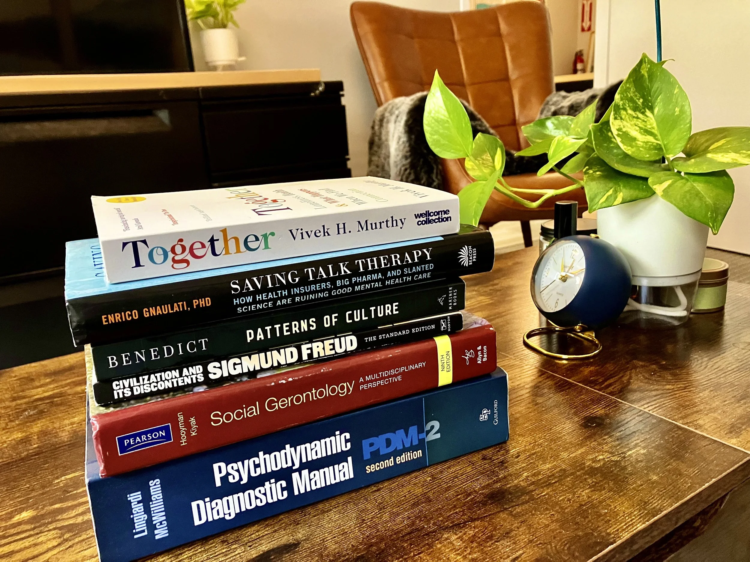 A stack of academic textbooks on psychology, social gerontology, and cultural studies placed on a wooden table, with a plant, a clock, and some other objects in the background.