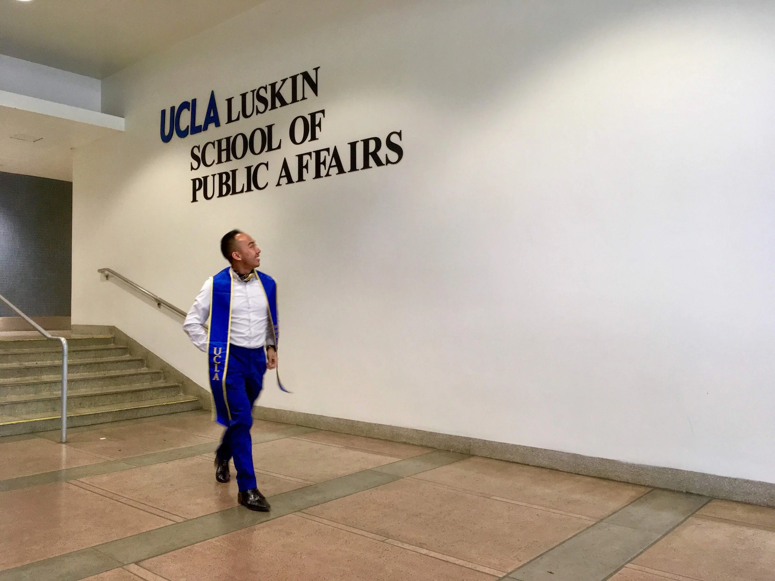 A man dressed in blue academic regalia walking up a staircase at UCLA Luskin School of Public Affairs.
