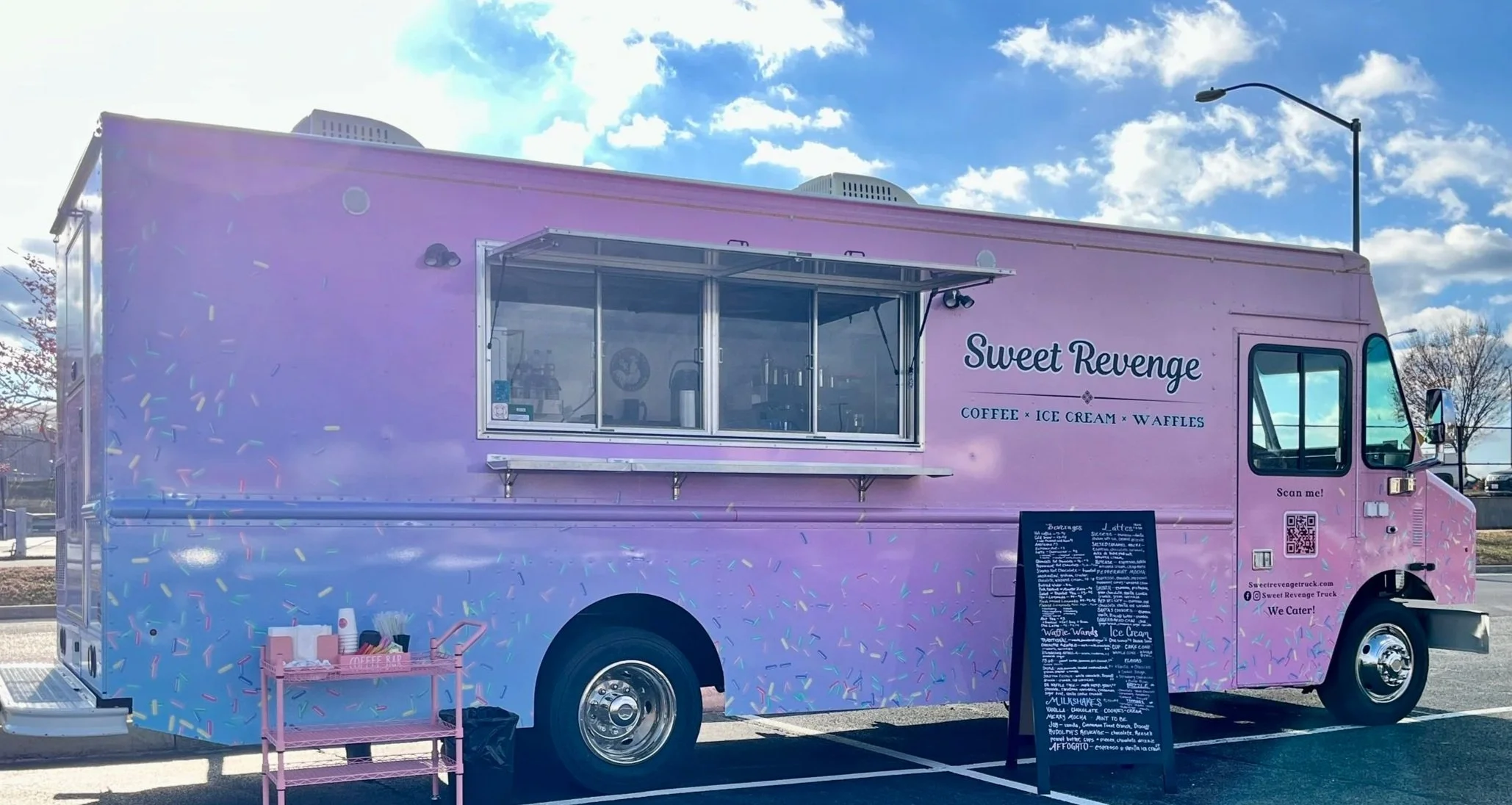 A pink food truck called Sweet Revenge offering coffee, ice cream, and waffles, parked in a lot on a sunny day with a blue sky and scattered clouds, with a chalkboard menu outside.
