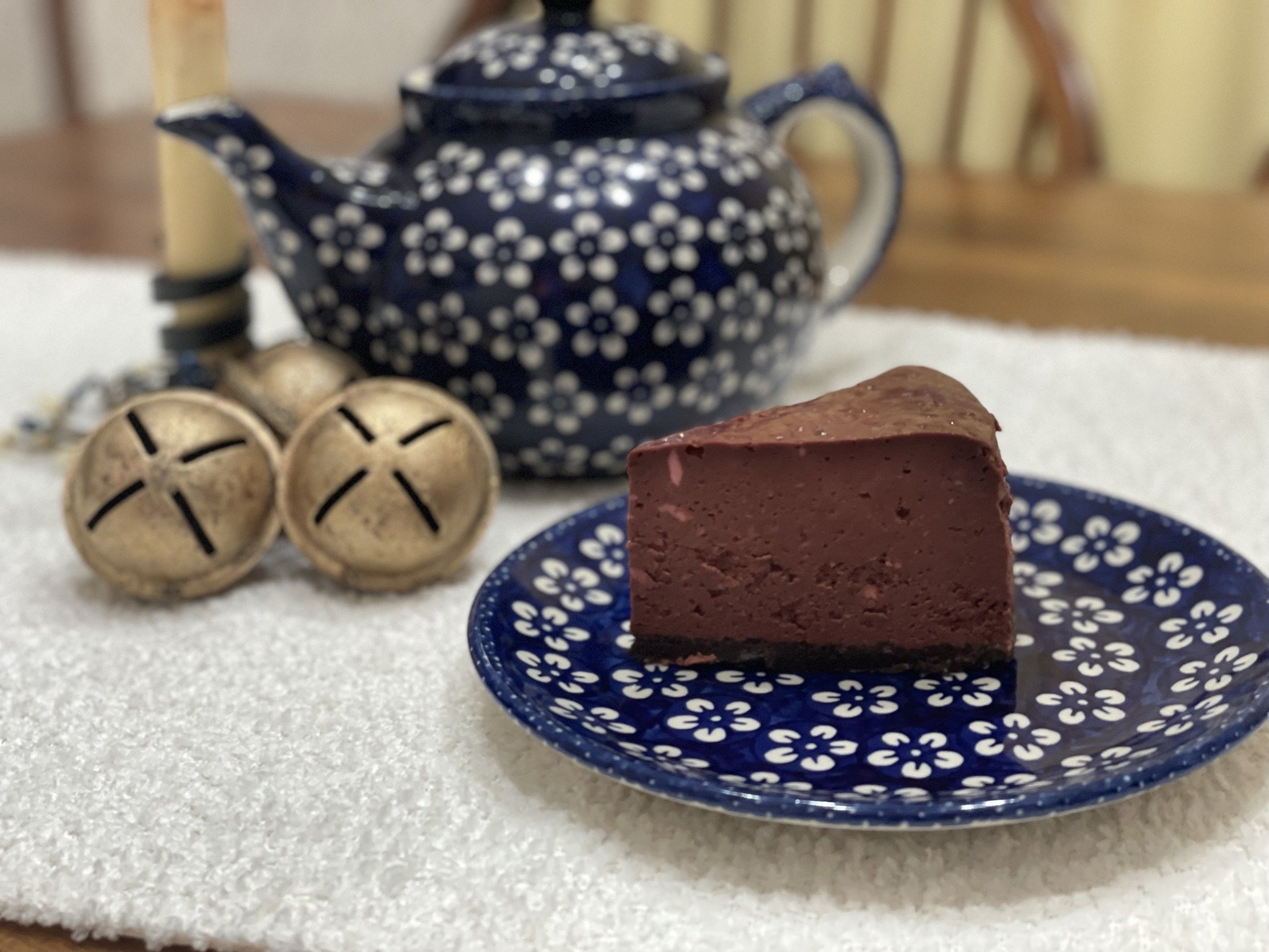 A slice of red velvet cake on a blue plate with white floral pattern, a blue teapot with white floral pattern, and two brass bells on a white tablecloth.