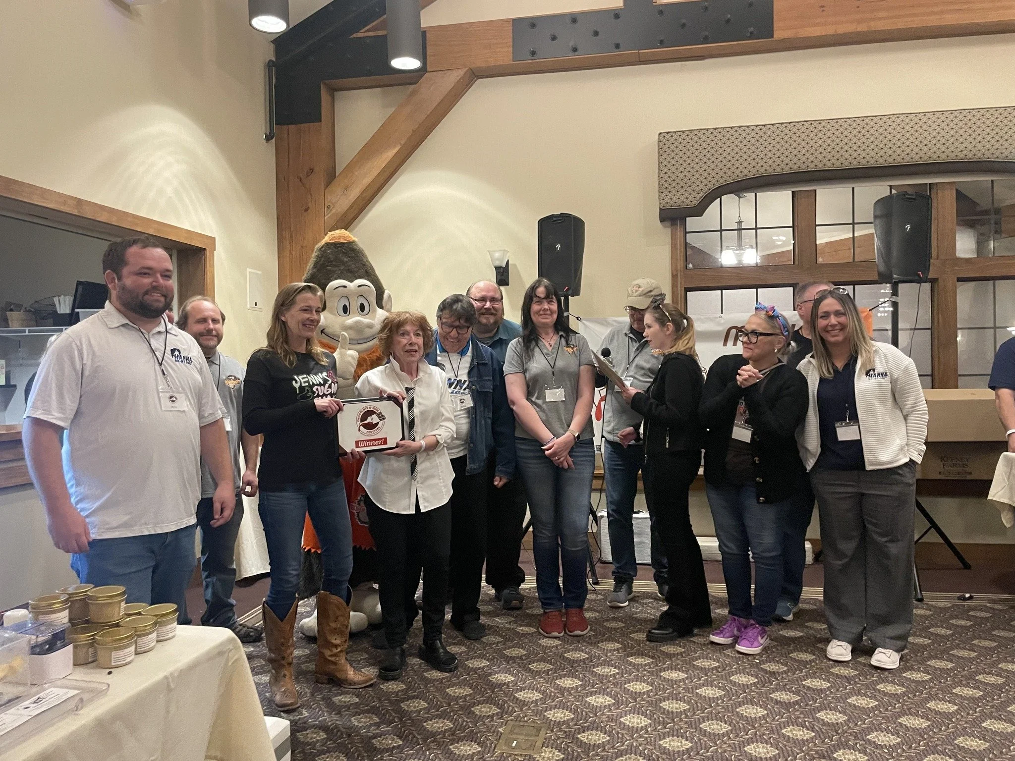 Group of people celebrating indoors, with a person in a mascot costume, one person holding a prize, and others clapping and smiling.