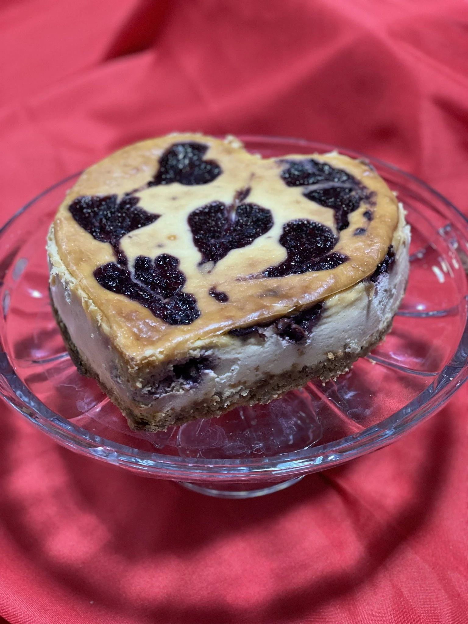 Heart-shaped cheesecake with blueberry swirls on top, served on a glass cake stand, against a red fabric background.