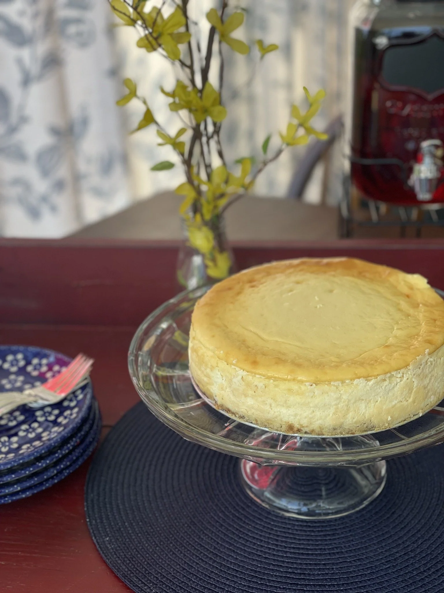 A cheesecake on a glass cake stand on a black placemat, with blue and white stacked plates and a fork to the left. A vase with yellow-green leafy branches is in the background on a wooden table.