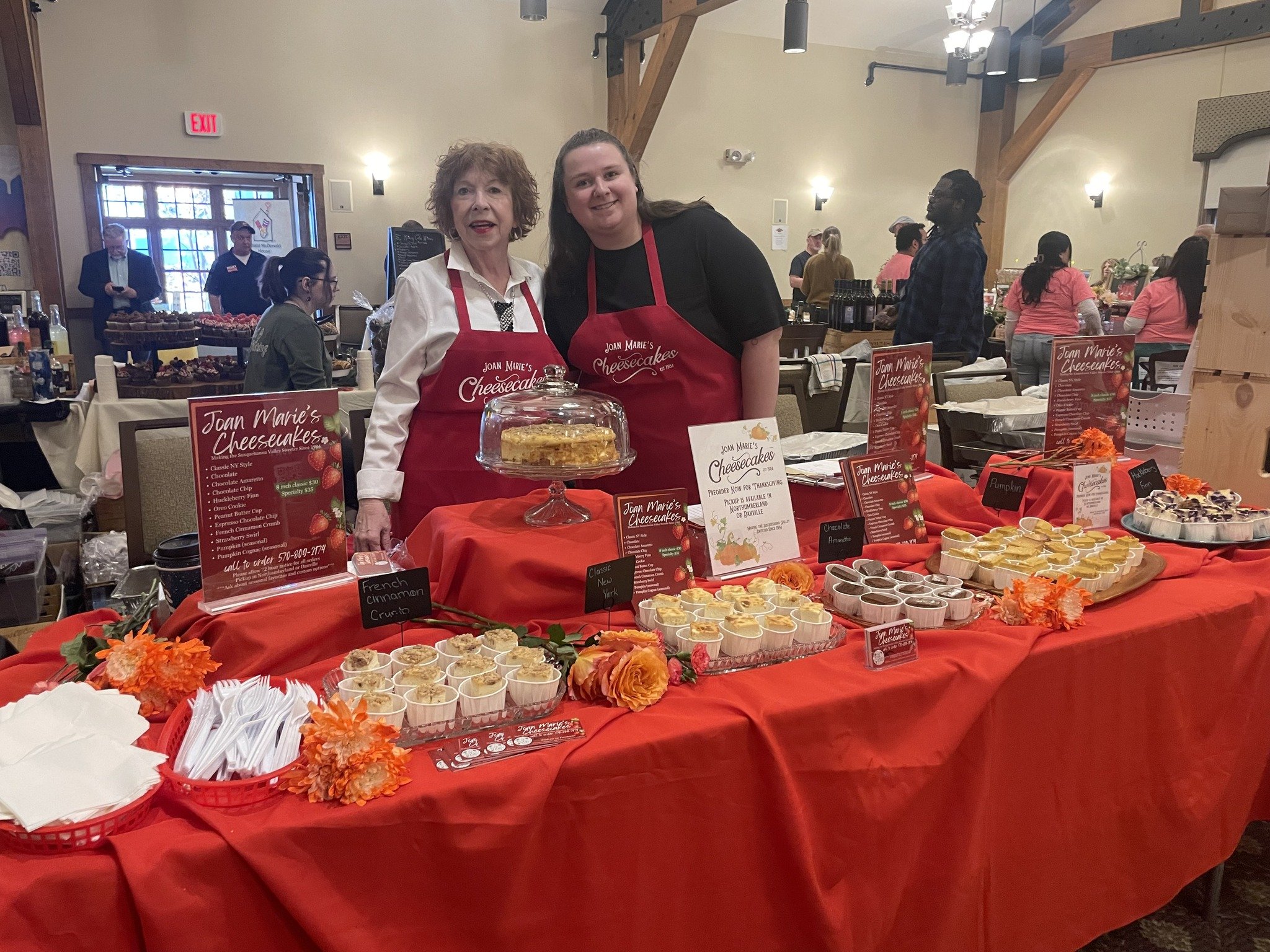 Two women - one Joan Marie - wearing red aprons behind a table with cheesecakes at an indoor event. The table has a red tablecloth, orange flowers, and signs listing cheesecake flavors. Other people and tables are visible in the background.