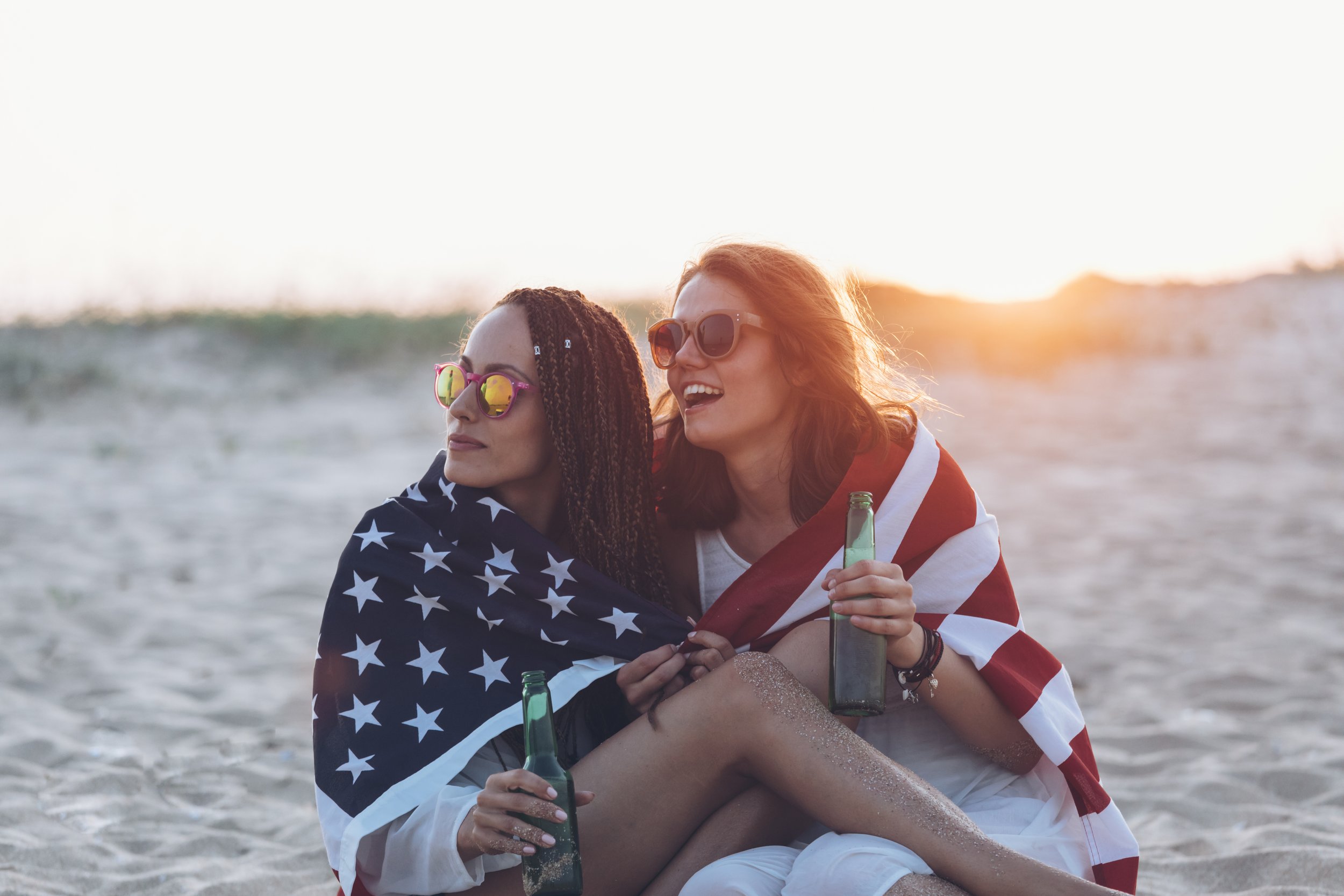 Two women sitting on a sandy beach, wrapped in an American flag, holding beer bottles and smiling, with sunglasses on.