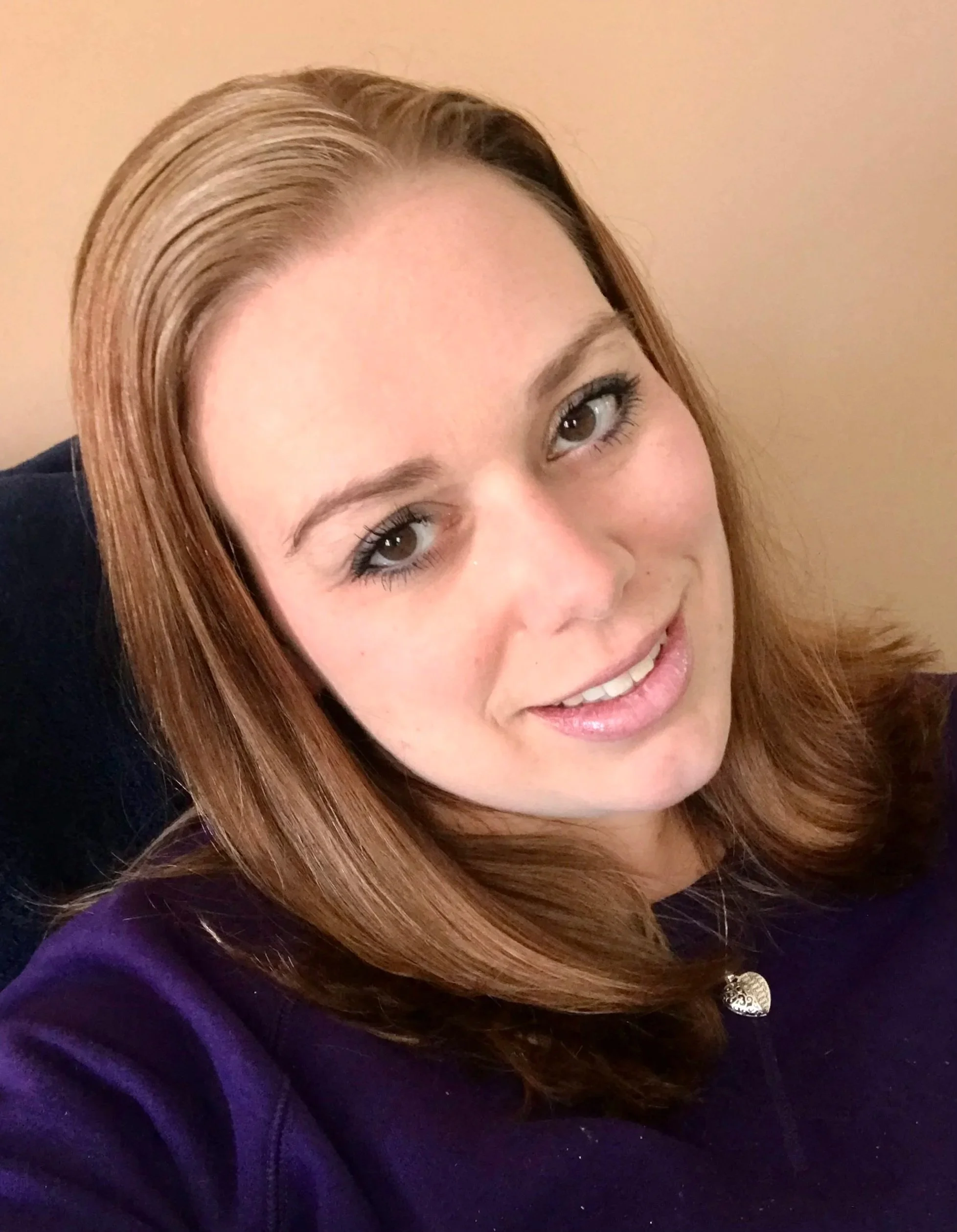 A woman with shoulder-length light brown hair, hazel eyes, and light skin, smiling and looking at the camera, wearing a purple top with a decorative button.