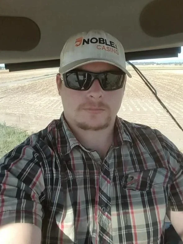 A man taking a selfie inside a vehicle, wearing a beige cap with a "Noble Casino" logo, dark sunglasses, and a plaid shirt. Outside the vehicle, there are fields with dirt and a clear sky.