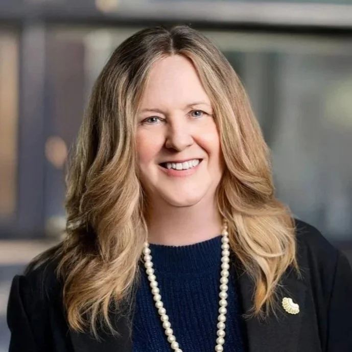 Smiling person with long hair wearing a blue shirt and black blazer in an indoor setting.