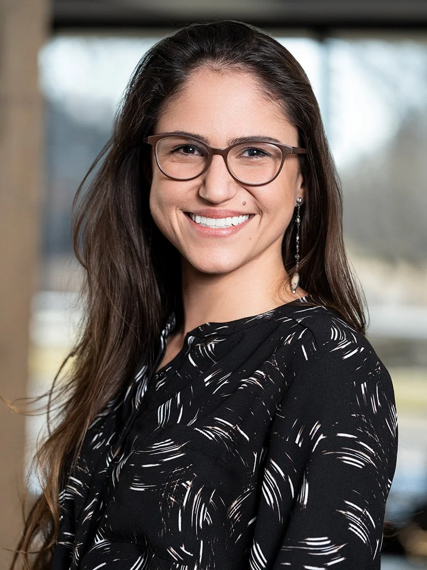 Woman with glasses smiling, wearing a black patterned shirt, standing indoors.