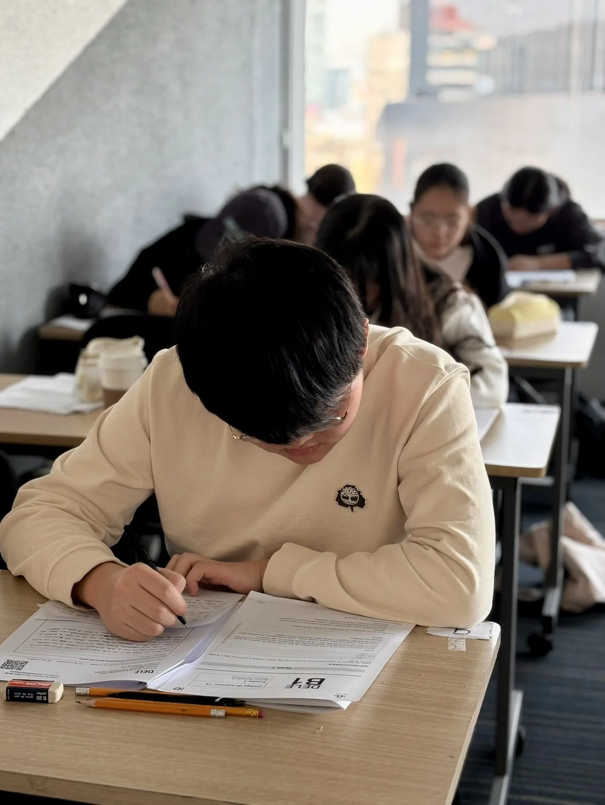 Estudiante concentrado en un examen en un aula con otros jóvenes estudiando en el fondo.