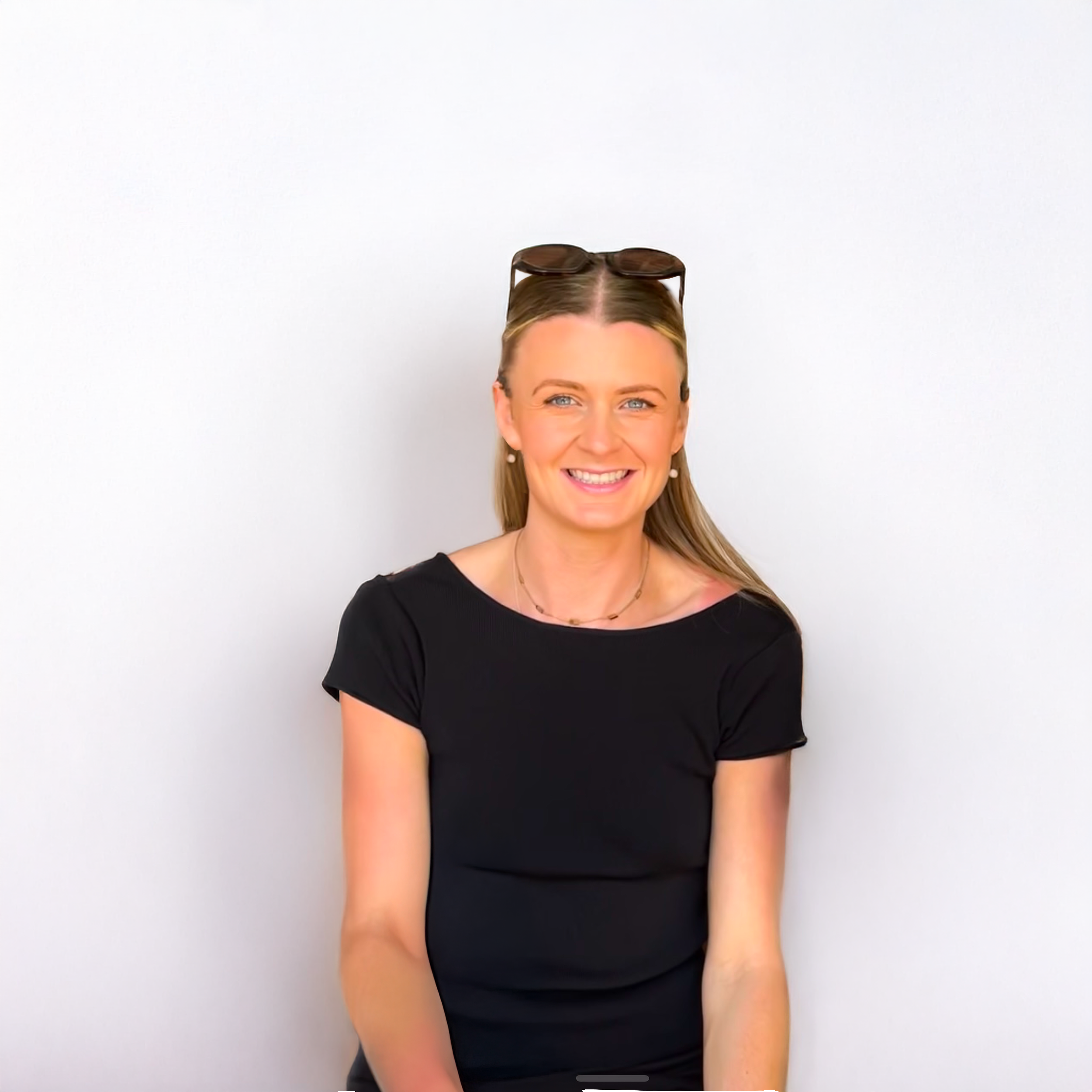 A smiling woman with blonde hair and blue eyes, wearing a black top and sunglasses on her head, sitting against a plain white background.