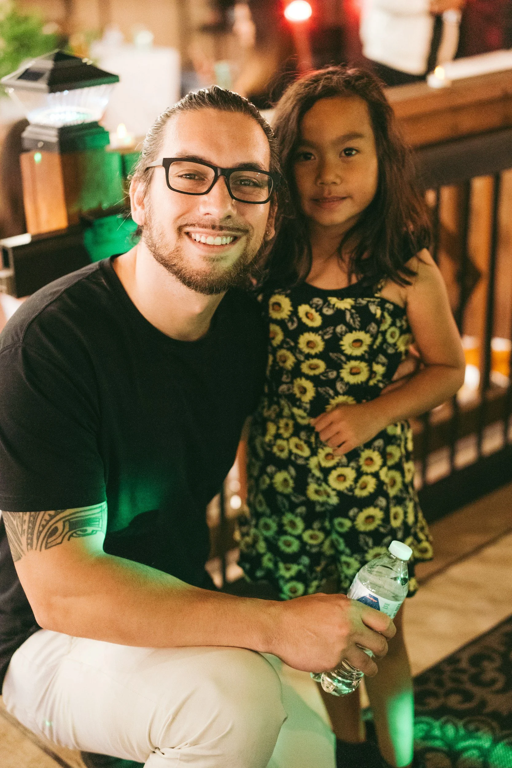 A smiling man with glasses and a tattoo on his arm sitting next to a young girl in a floral dress at a lively indoor gathering.