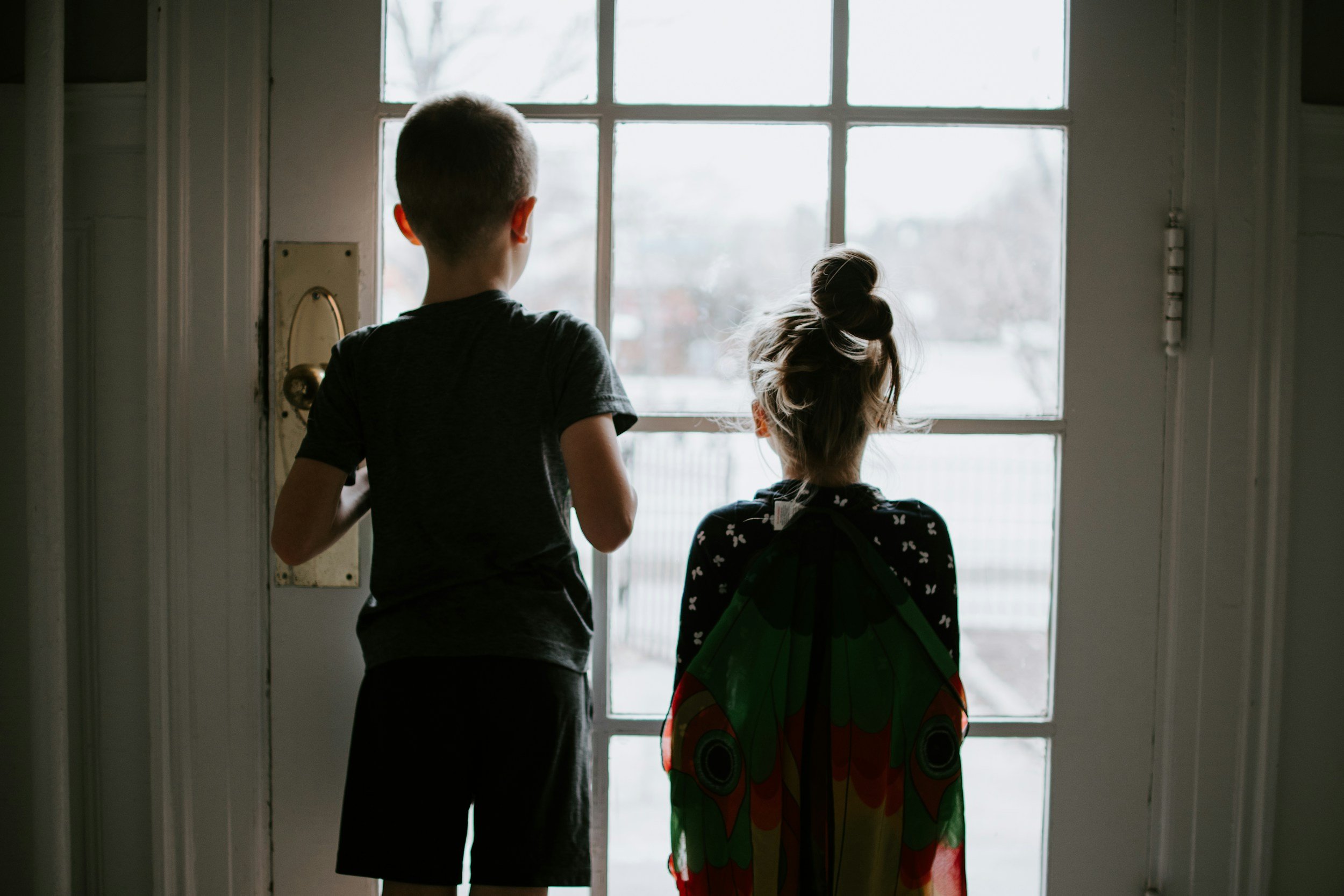 A boy and a girl stand at a glass door, looking outside on a snowy day. The girl is wearing butterfly wings. It's daytime.