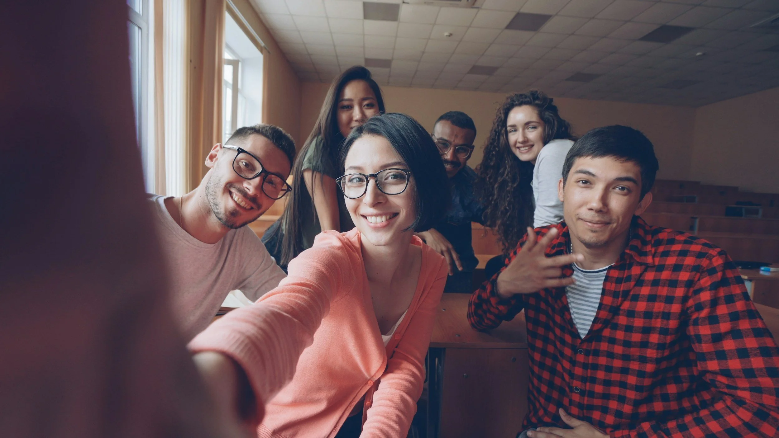 Group of six young adults smiling and taking a selfie in a classroom with wooden desks and large windows.