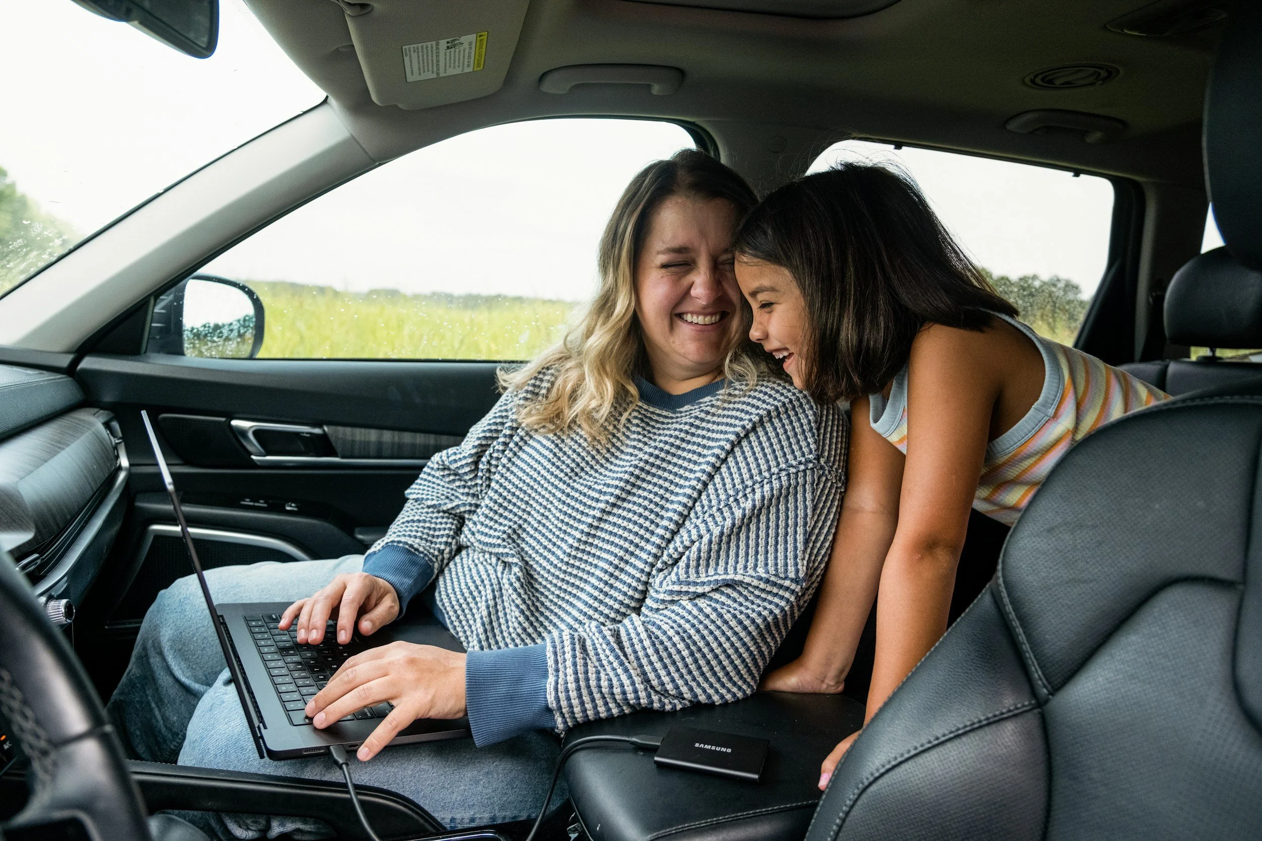 A woman and a girl laughing together inside a car, with the woman sitting in the passenger seat and the girl leaning over from the back seat. The woman is using a laptop on her lap.